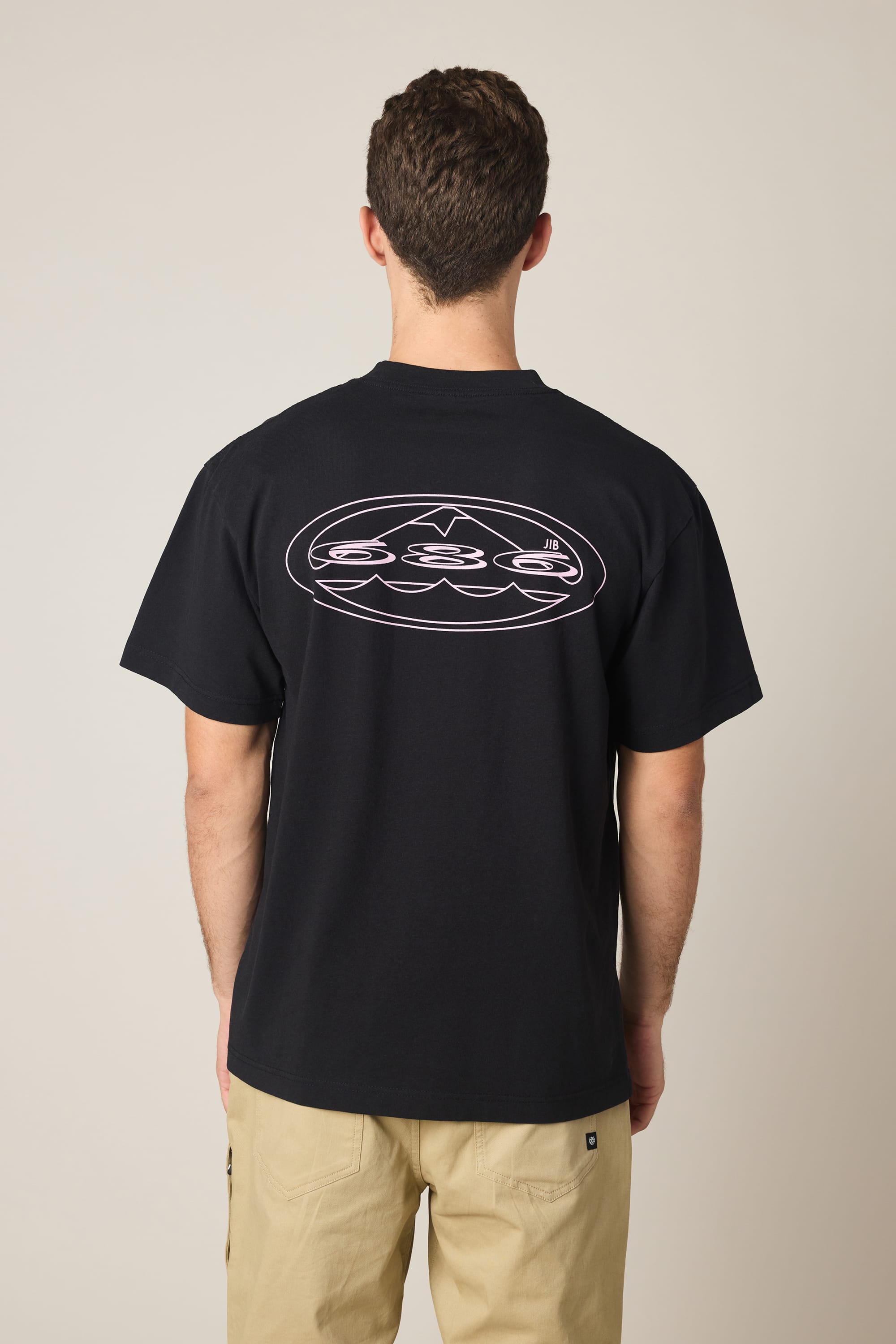 A man wears the 686 Men's Jib Relaxed SS Tee in black, featuring a white-outlined back graphic. He pairs it with light beige pants, standing with his back to the camera against a plain, neutral background.