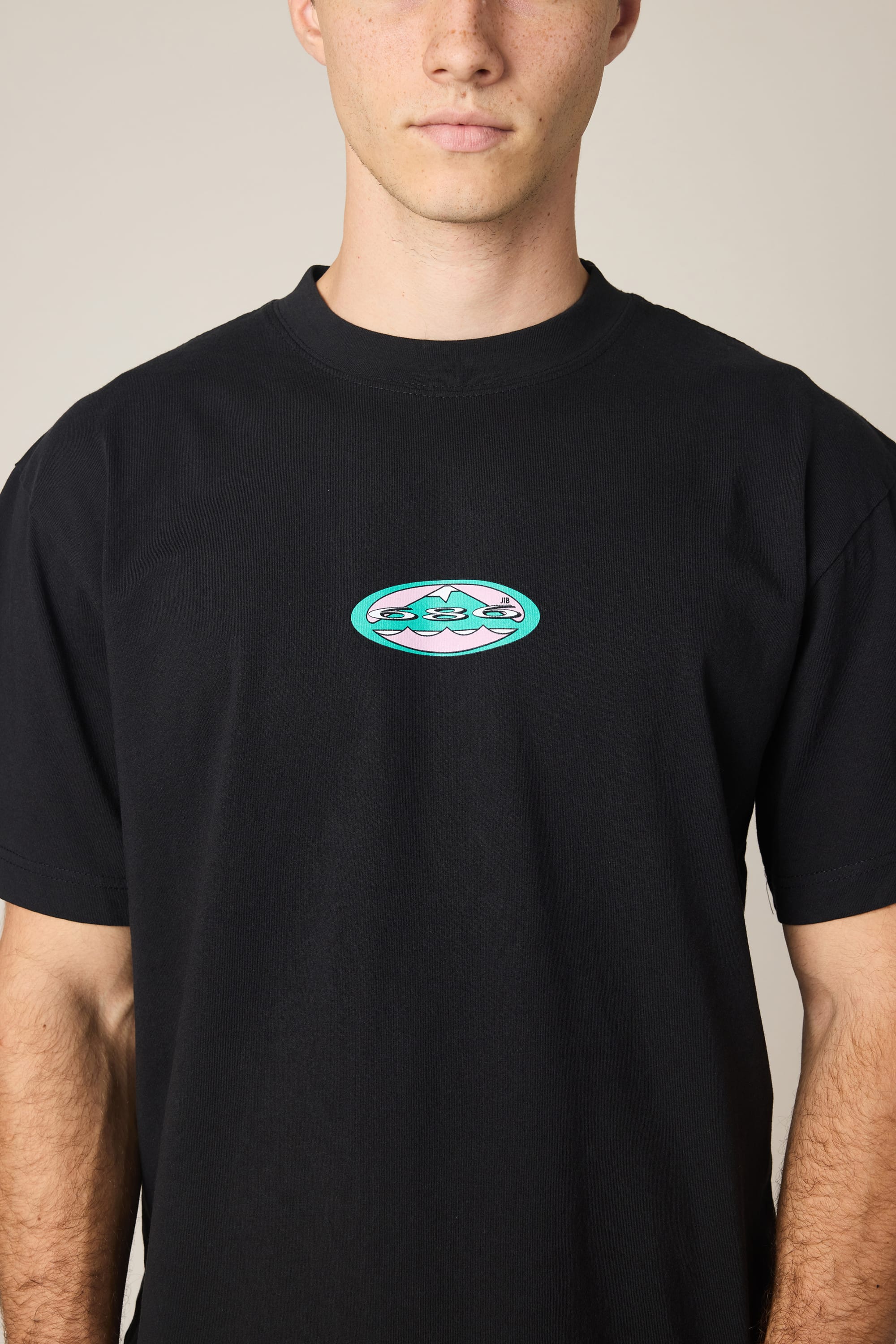 A person wearing the 686 Men's Jib Relaxed SS Tee by 686, a black 100% cotton t-shirt featuring a small, colorful oval graphic on the chest, stands against a plain background.