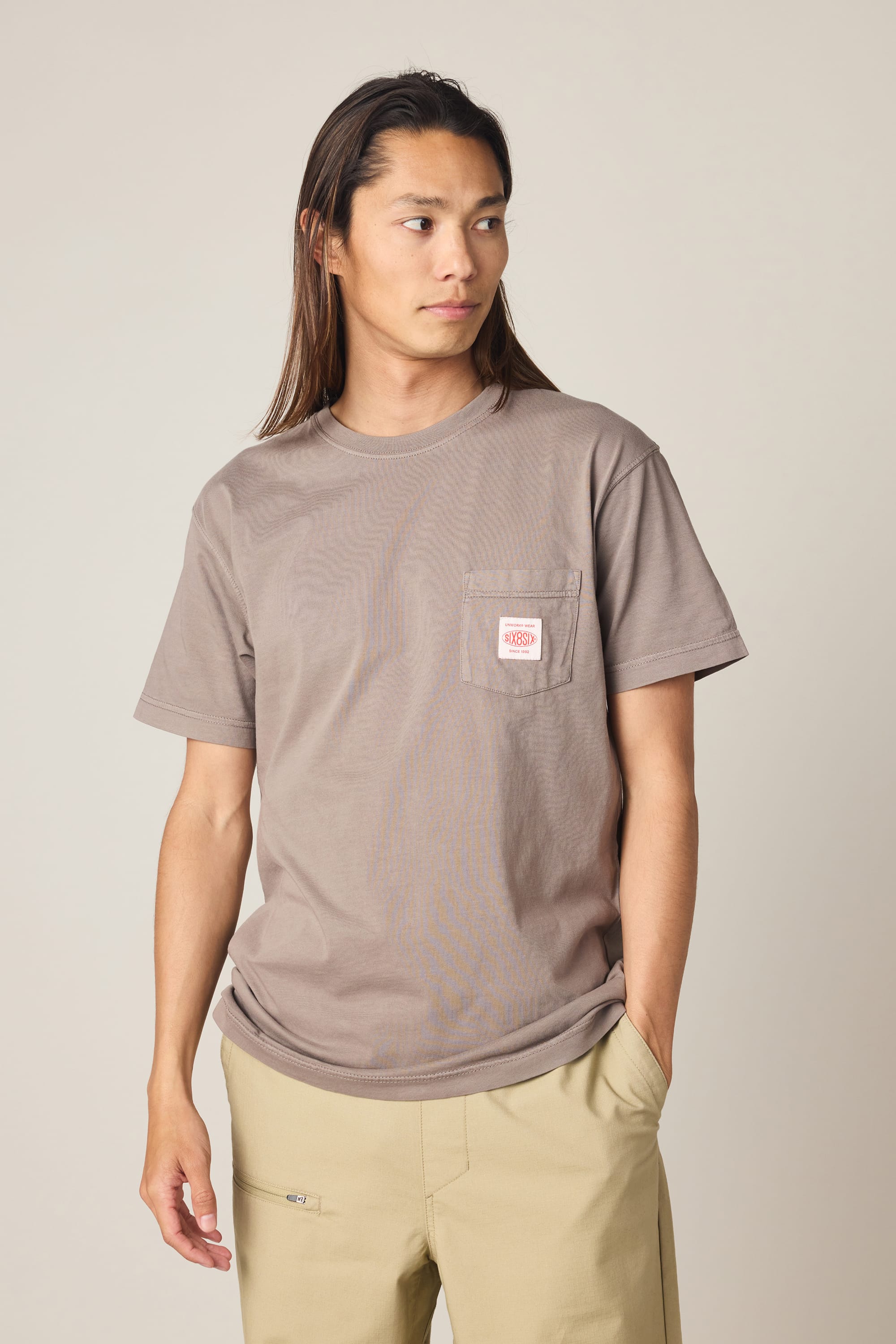 A person with long hair wears the 686 Men's Unwork Authentic SS Pocket Tee in taupe—crafted from 100% cotton and detailed with a small white and red patch on the chest pocket—styled with light beige pants and standing against a plain light background.