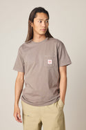 A person with long brown hair models the 686 Men's Unwork Authentic SS Pocket Tee in taupe with a chest pocket, paired with beige pants, standing against a plain background and looking to the side with one hand in their pocket.
