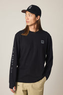 A person with long hair wears a black cap, beige pants, and the 686 Men's Let's Chill Waffle LS Crew by 686, standing with one hand in a pocket and looking to the side against a neutral background.