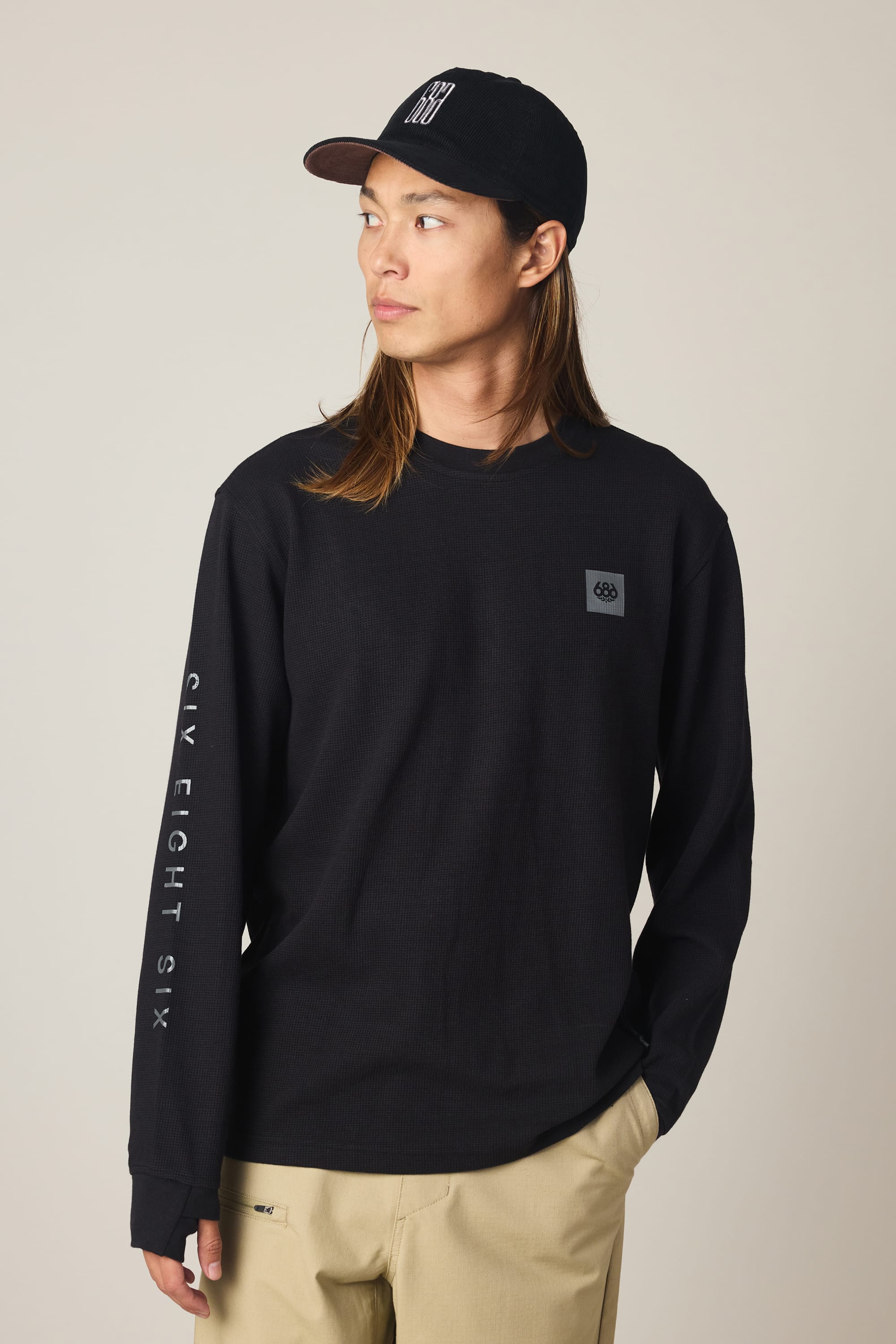 A person with long hair wears a black cap, beige pants, and the 686 Men's Let's Chill Waffle LS Crew by 686, standing with one hand in a pocket and looking to the side against a neutral background.