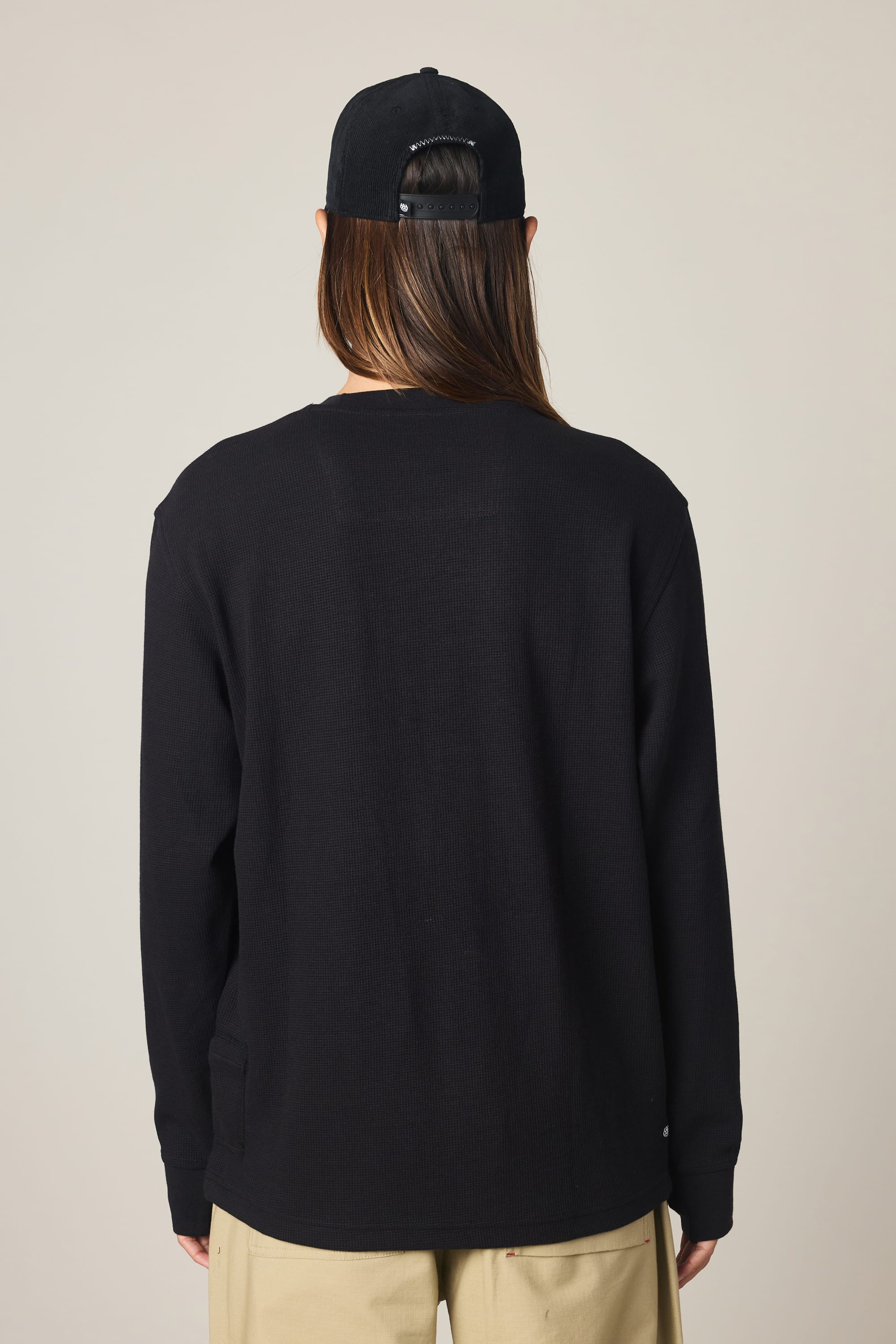 A man is shown from the back against a plain light background, wearing the 686 Men's Let's Chill Waffle LS Crew in black, paired with beige pants and a black cap.