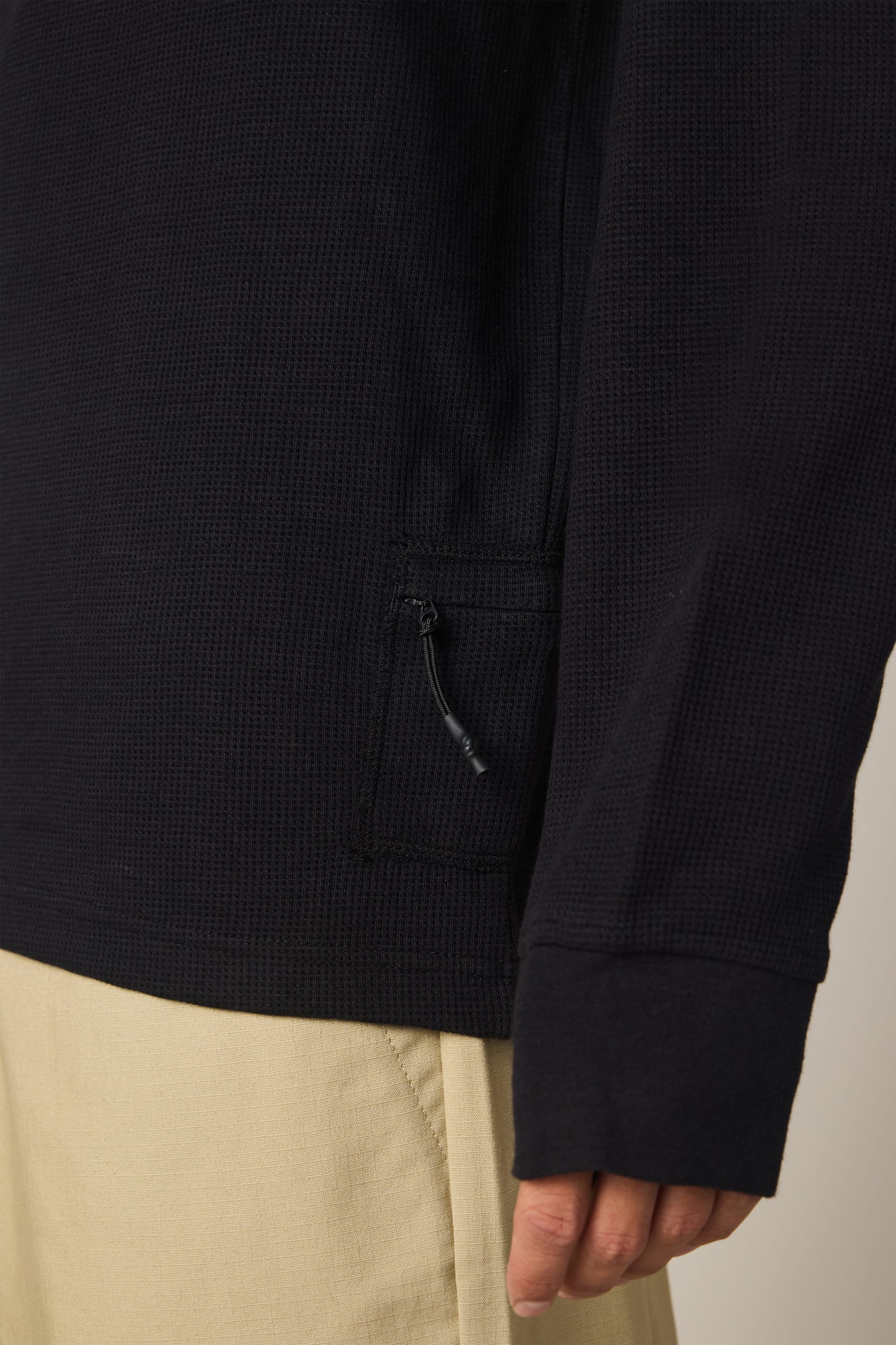 A close-up of a person wearing the 686 Men's Let's Chill Waffle LS Crew in black—a textured long-sleeve polyester t-shirt with a small zippered pocket—paired with light beige pants. Only the lower half of the shirt and a hand are visible.