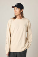 A person with long hair wears a beige 686 Men's Let's Chill Waffle LS Crew with a small chest logo and a black baseball cap, looking to the side with one hand in their pocket against a plain background.