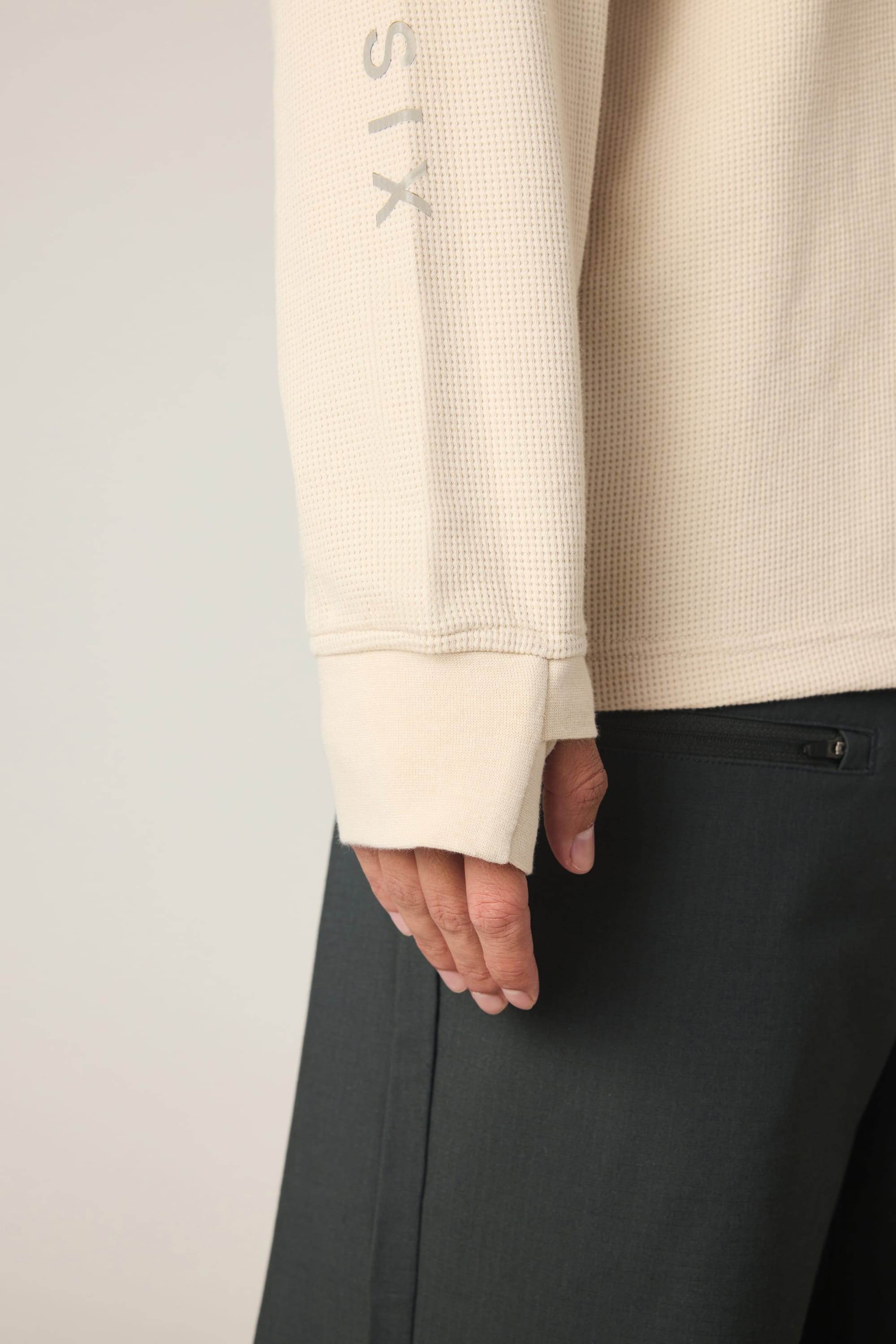 A person is wearing the 686 Men's Let's Chill Waffle LS Crew by 686, a cream long-sleeve textured top resembling a men's polyester t-shirt, with the sleeve partially covering their hand. They are also dressed in dark-colored pants with a visible back pocket, against a plain light-colored background.
