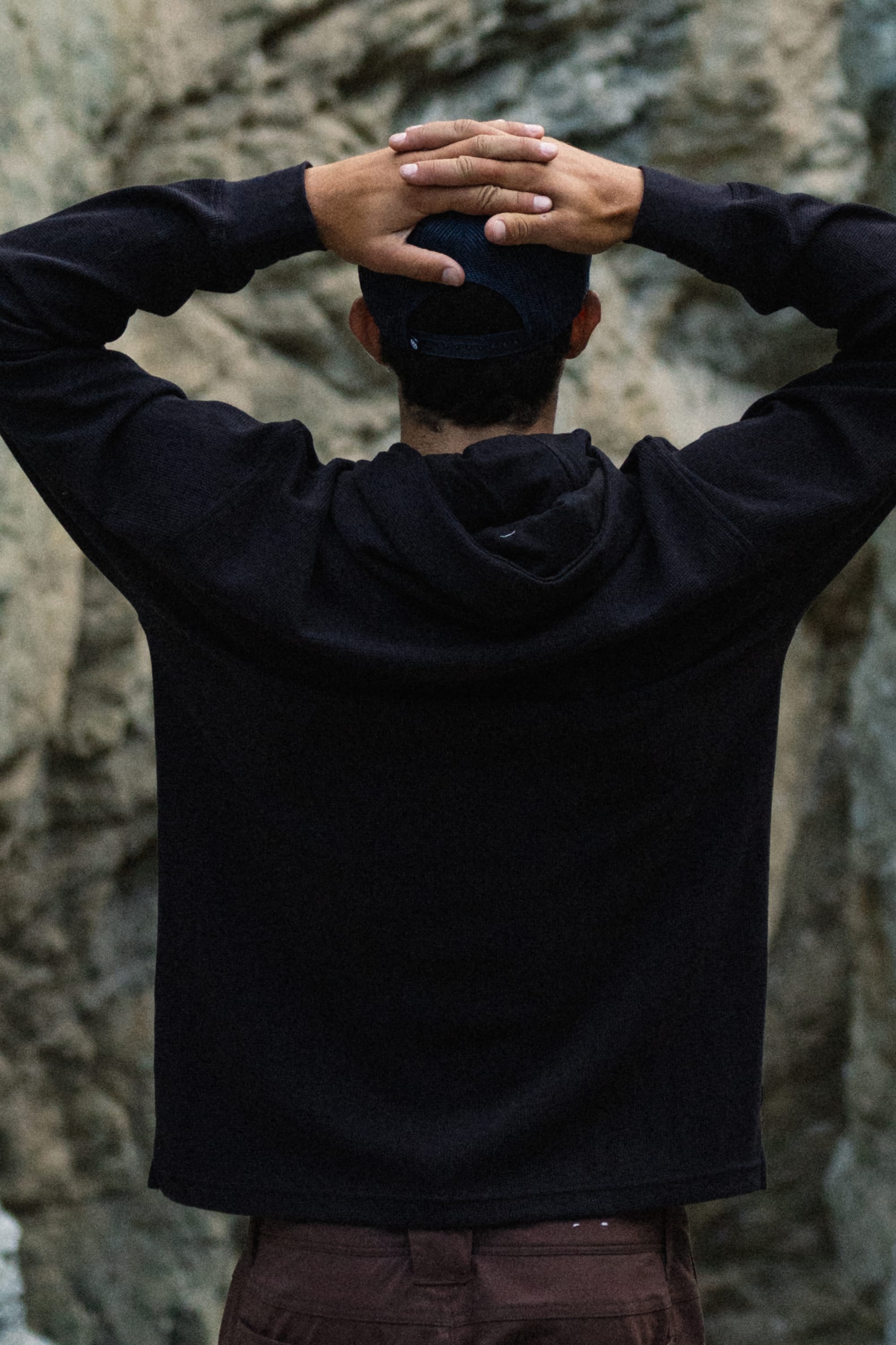Wearing the 686 Men's Let's Chill Waffle Hoody and a cap over a men's polyester t-shirt, an individual stands with their back to the camera, hands clasped on their head, against a backdrop of rugged rocks.