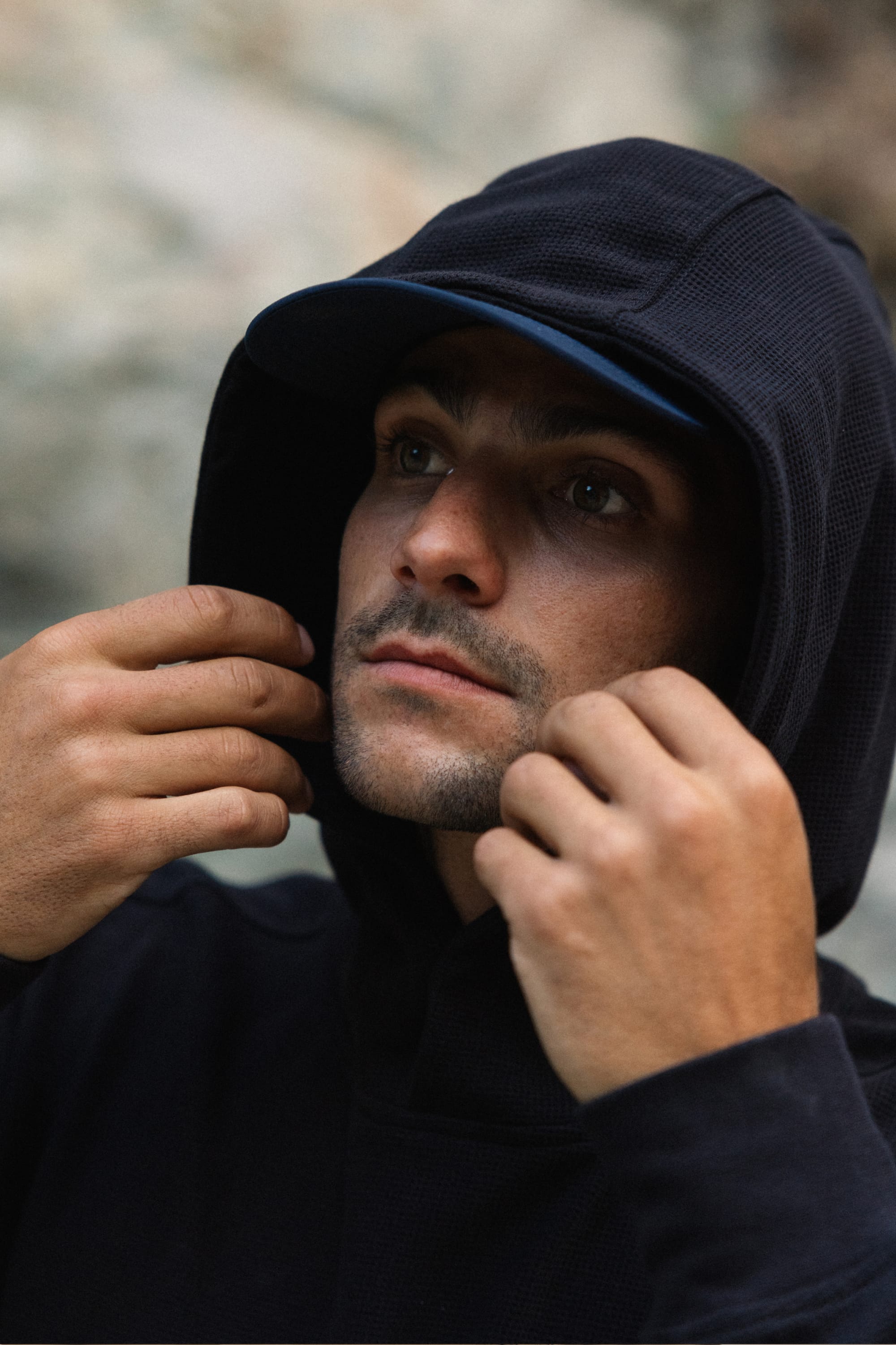 A person wearing the 686 Men's Let's Chill Waffle Hoody by 686 over a men's polyester t-shirt and cap looks up while adjusting the hood with both hands, with a blurred background that draws focus to their face and hands.