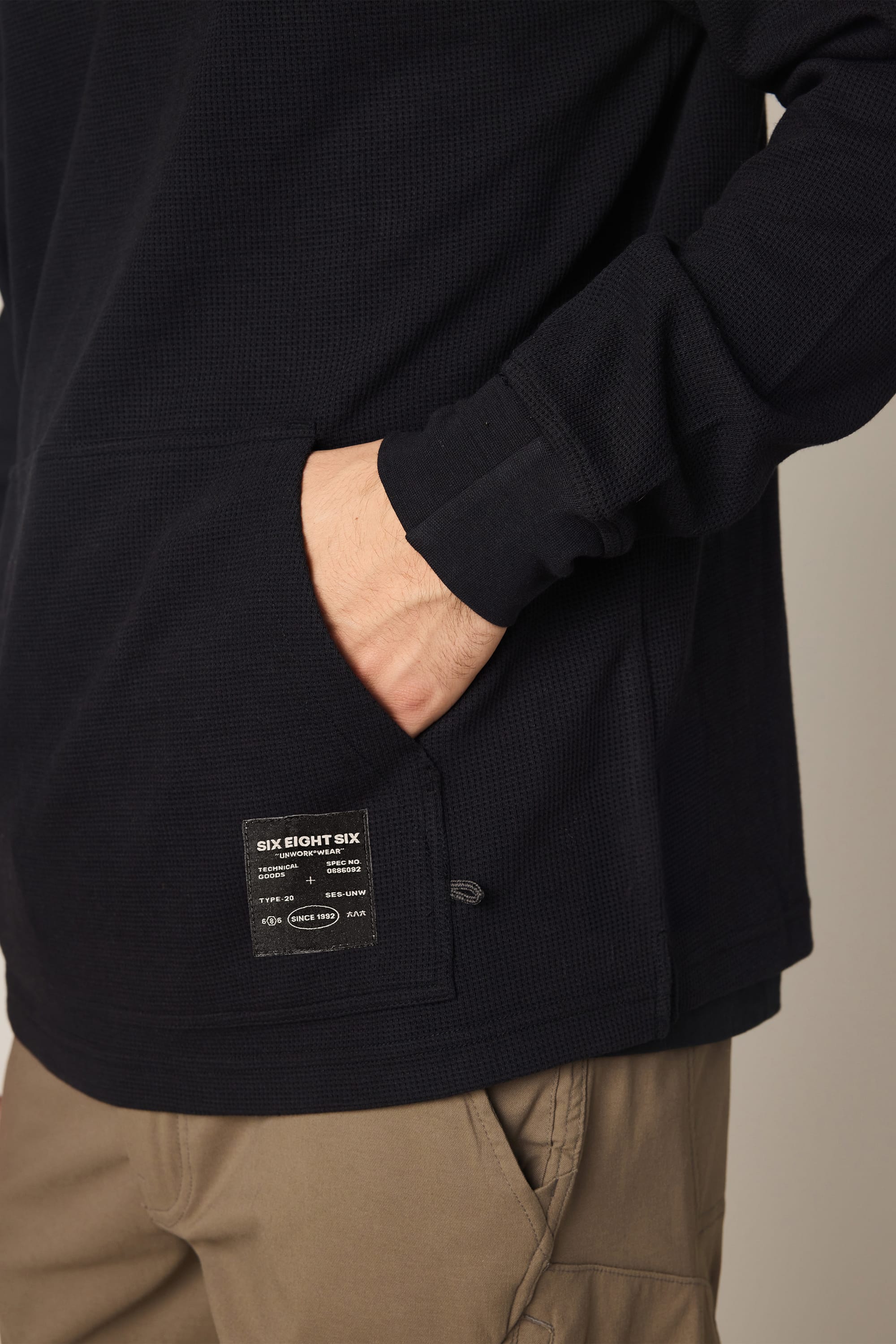 A person wears the 686 Men's Let's Chill Waffle Hoody in black with a patch near the pocket and tan pants, right hand in the front pocket. Only their torso, part of an arm, and pants are visible.