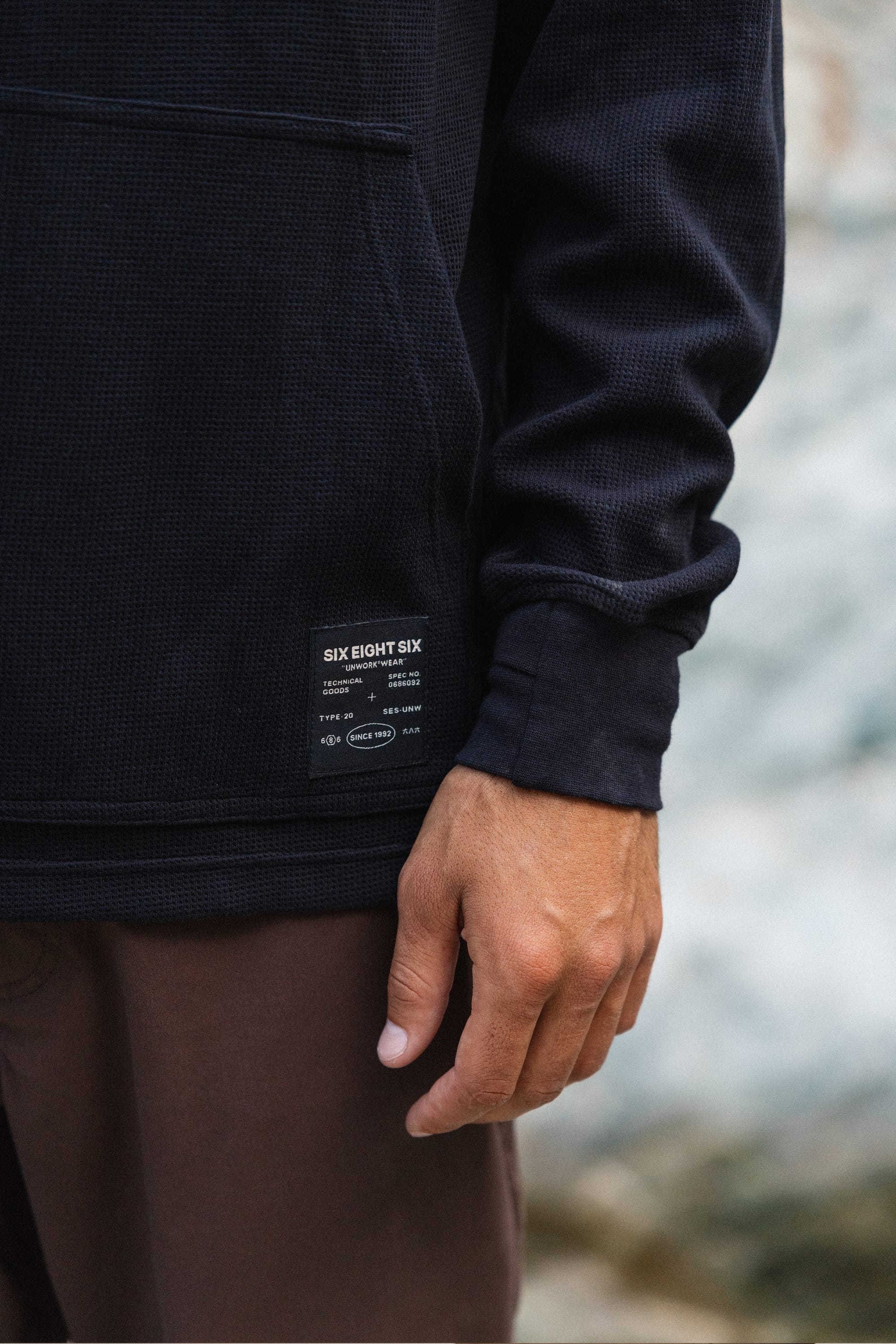 A person wearing the 686 Men's Let's Chill Waffle Hoody in black, featuring a visible 686 label, pairs it with brown pants while standing outdoors with one hand relaxed at their side—effortlessly layering the look as you might with a classic men's polyester t-shirt.