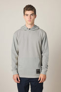 A young man with short brown hair stands against a plain light background wearing the 686 Men's Let's Chill Waffle Hoody from 686 over a men's t-shirt and dark pants. He has a neutral expression and a small black label near the hoodie’s hem.