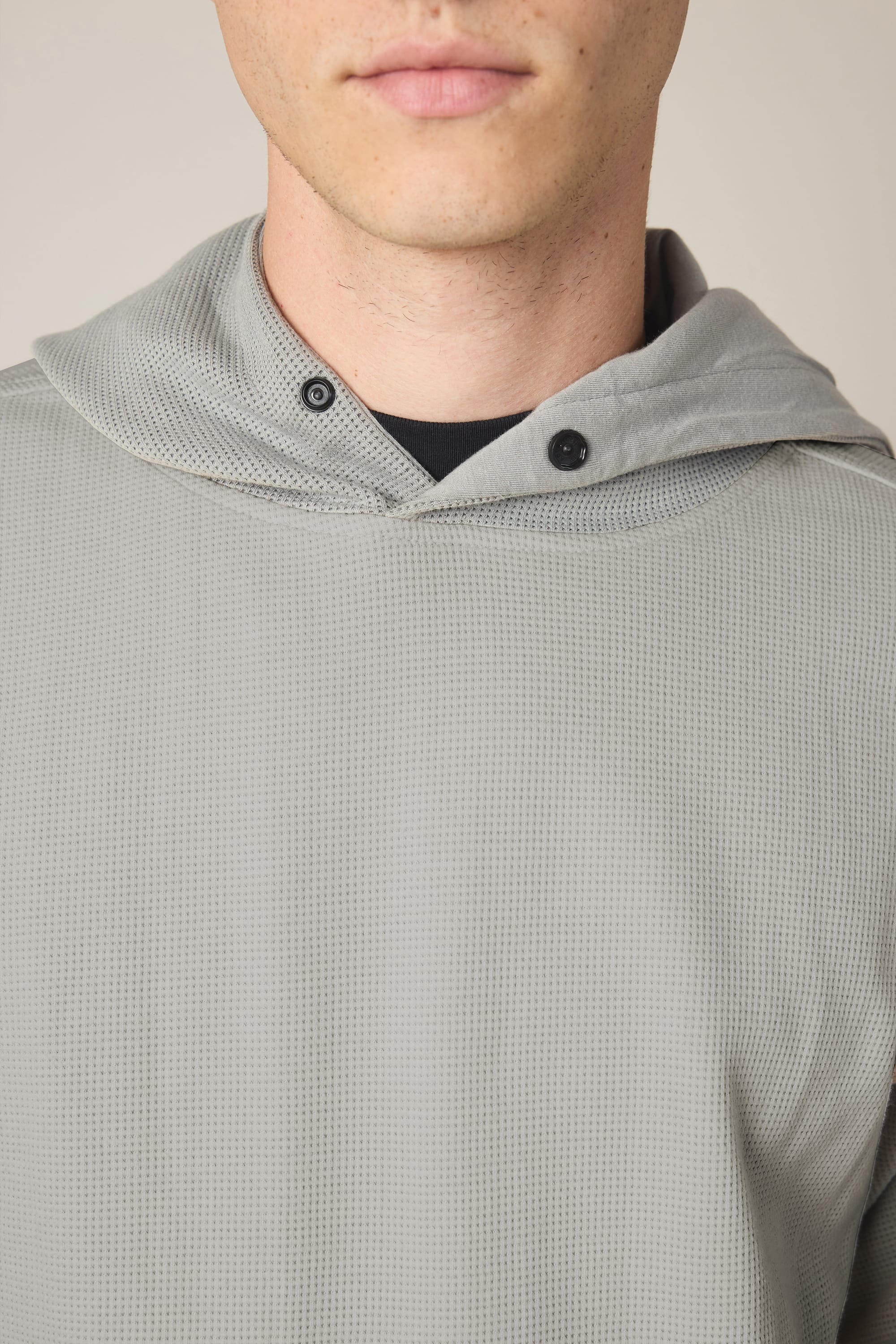 A close-up of a person wearing the 686 Men's Let's Chill Waffle Hoody in light gray, with only the lower half of their face visible. Layered underneath, a men’s polyester t-shirt subtly peeks out at the collar. The hoody from 686 features black snap buttons near the neck for added detail.