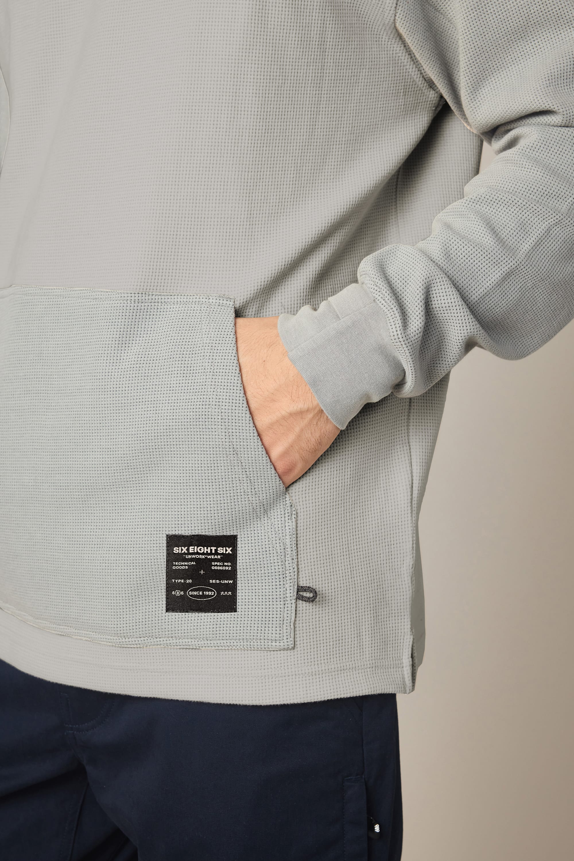 A person is wearing the 686 Men's Let's Chill Waffle Hoody—a light gray textured polyester hoodie with a front pocket and a visible black label—paired with dark pants, with their right hand partially tucked into the pocket.