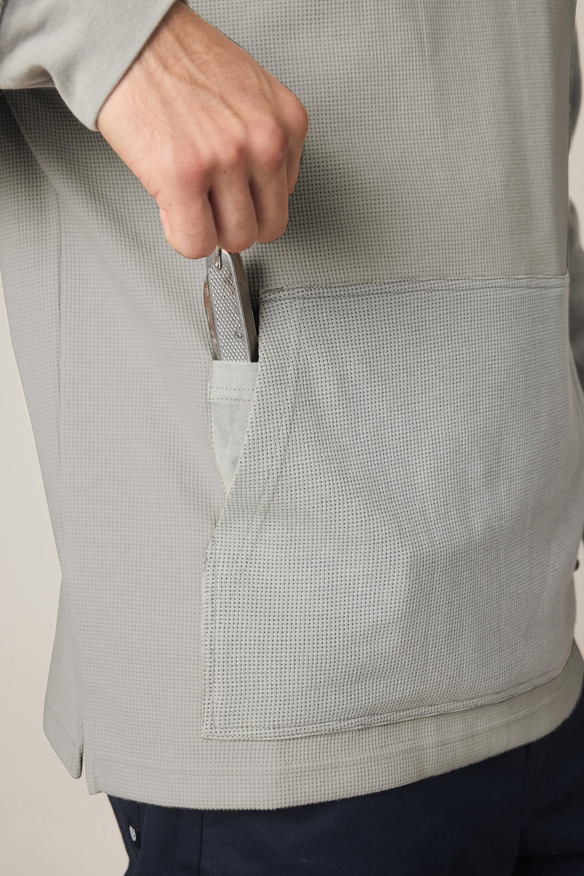A person wearing a textured light gray 686 Men's Let's Chill Waffle Hoody places a smartphone into the front pocket of the hoody. Only the hand, part of the arm, and torso are visible.