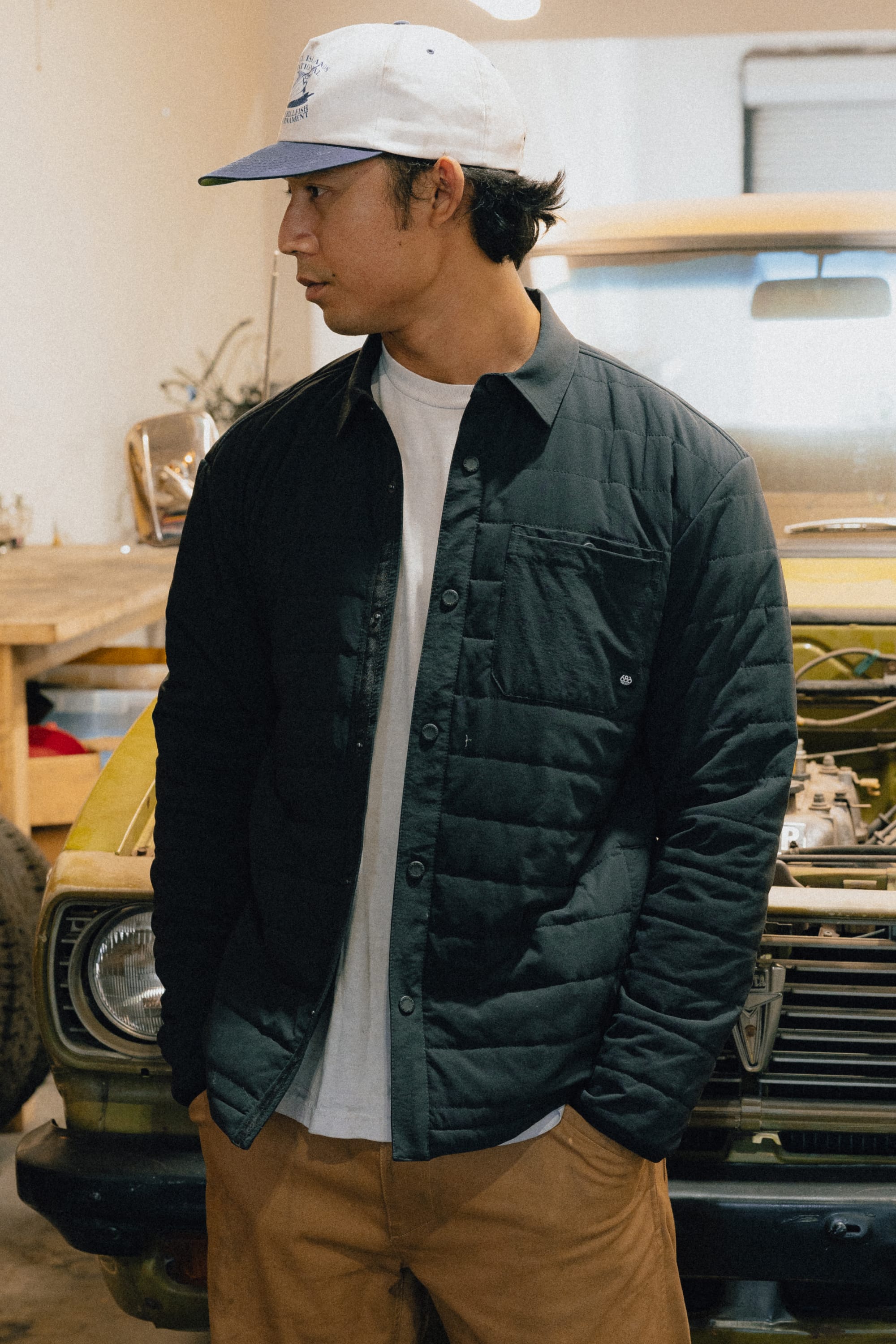 A man in a white cap, wearing the 686 Men's Thermaldry™ Insulated Shacket by 686 over a white shirt and brown pants, stands in front of an old yellow car in a garage, looking to his left.
