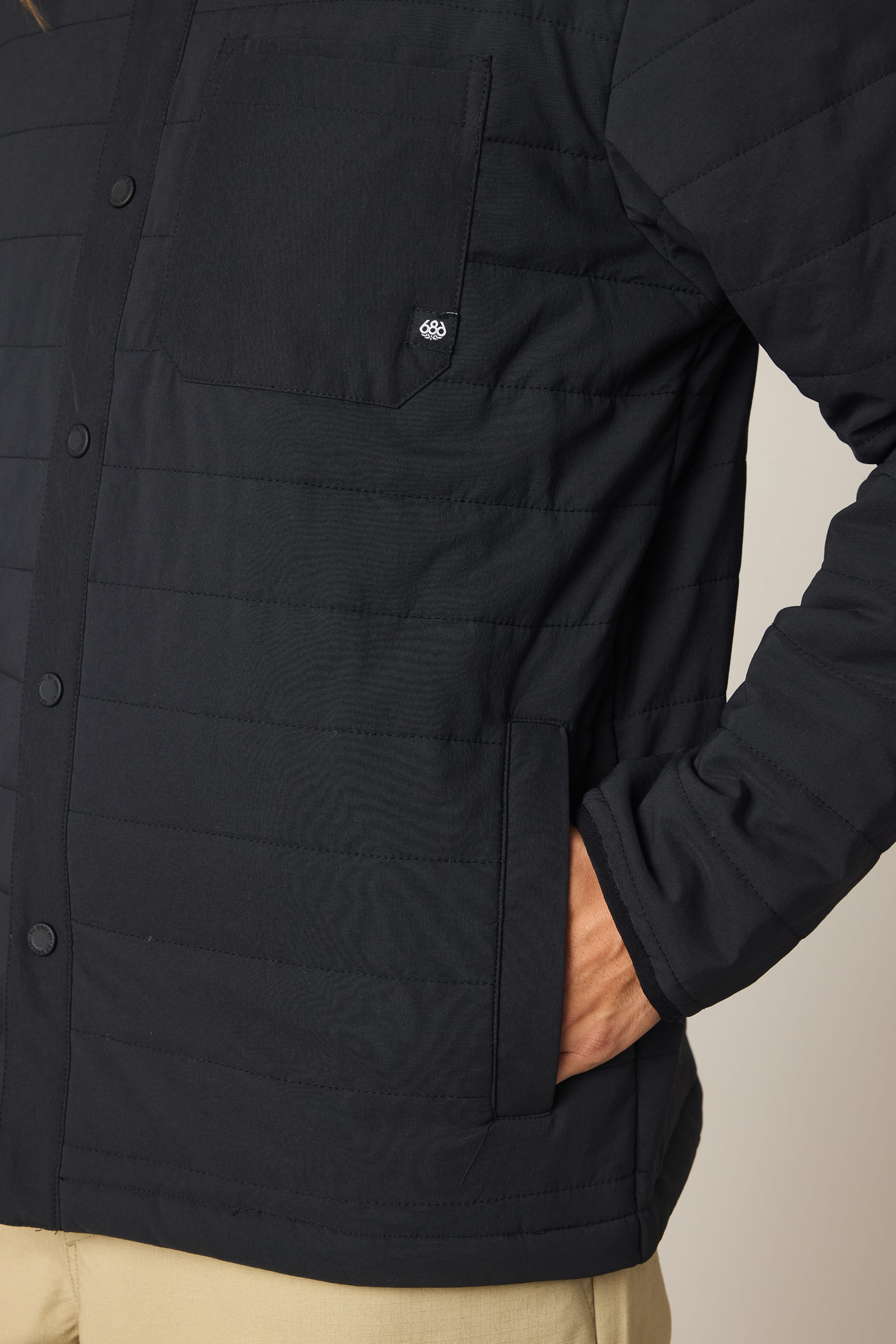 A man wears the 686 Men's Thermaldry Insulated Shacket—a black, quilted polyester jacket with snap buttons and a chest pocket featuring an embroidered logo—paired with beige pants, one hand tucked into a front pocket.