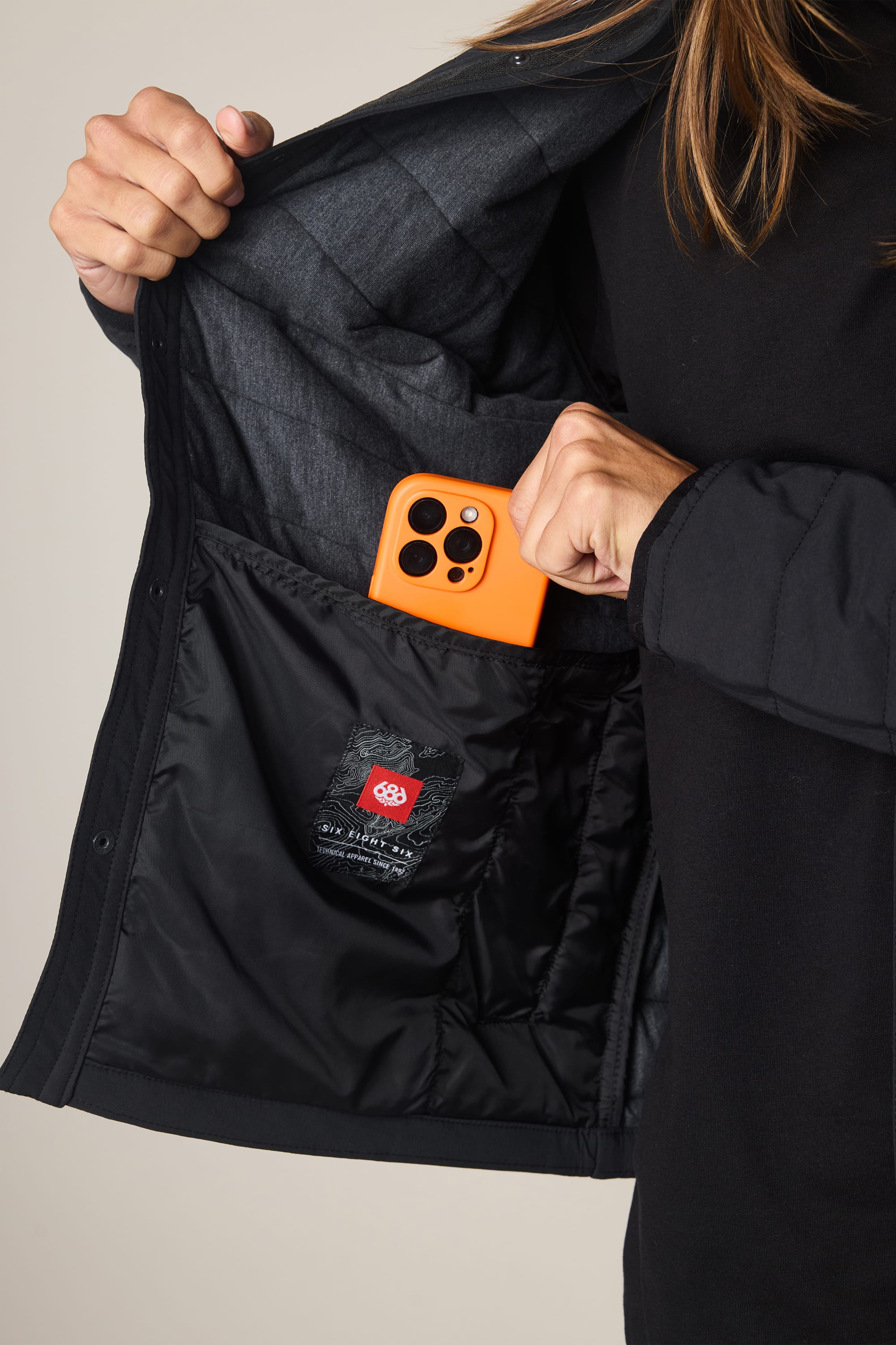 A person slips an orange smartphone into the inner pocket of a 686 Men's Thermaldry Insulated Shacket, showing the shacket's interior lining and the 686 label.