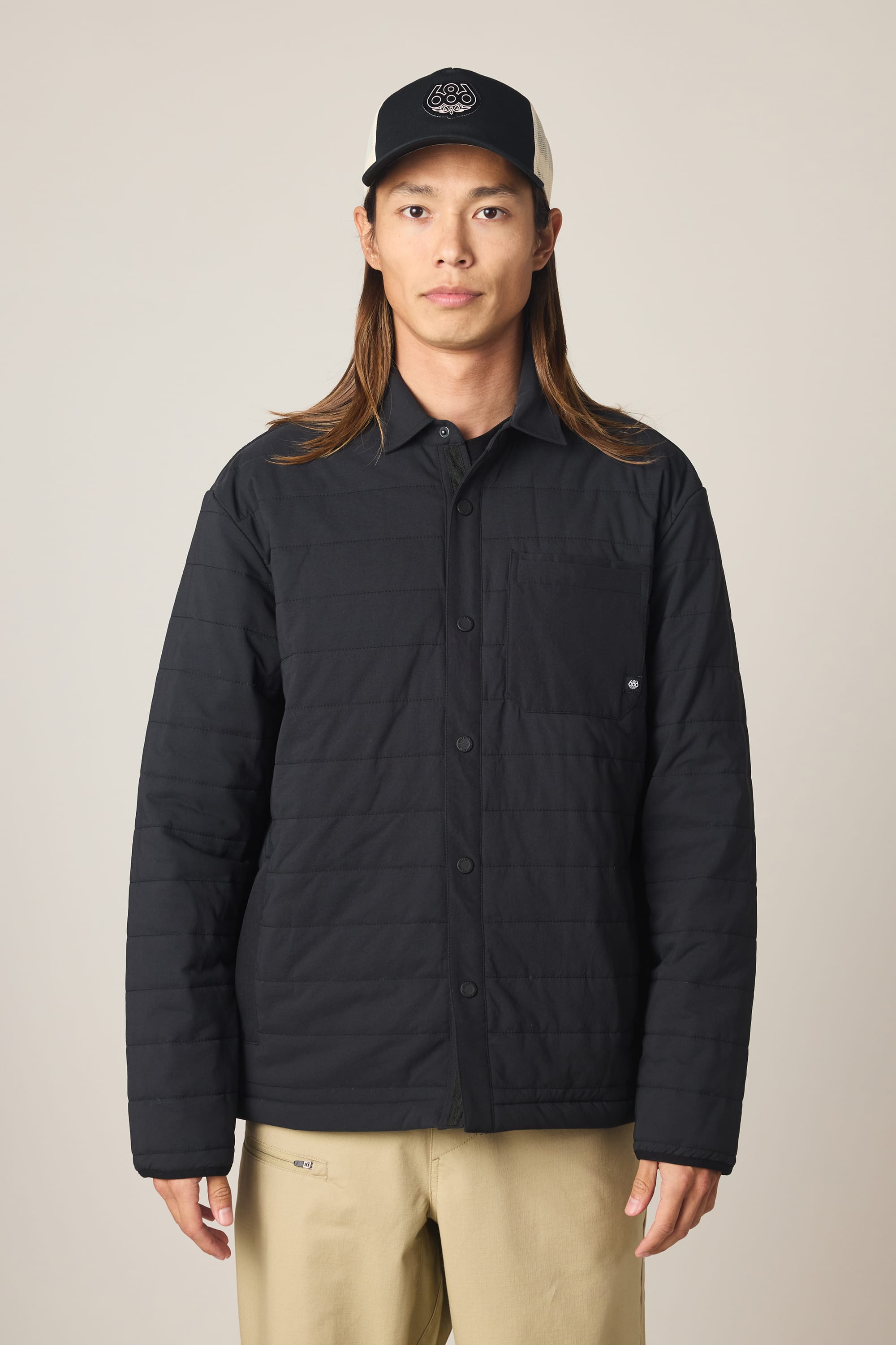 A person with long brown hair wears the 686 Men's Thermaldry Insulated Shacket, beige pants, and a black baseball cap, standing in front of a plain light background.