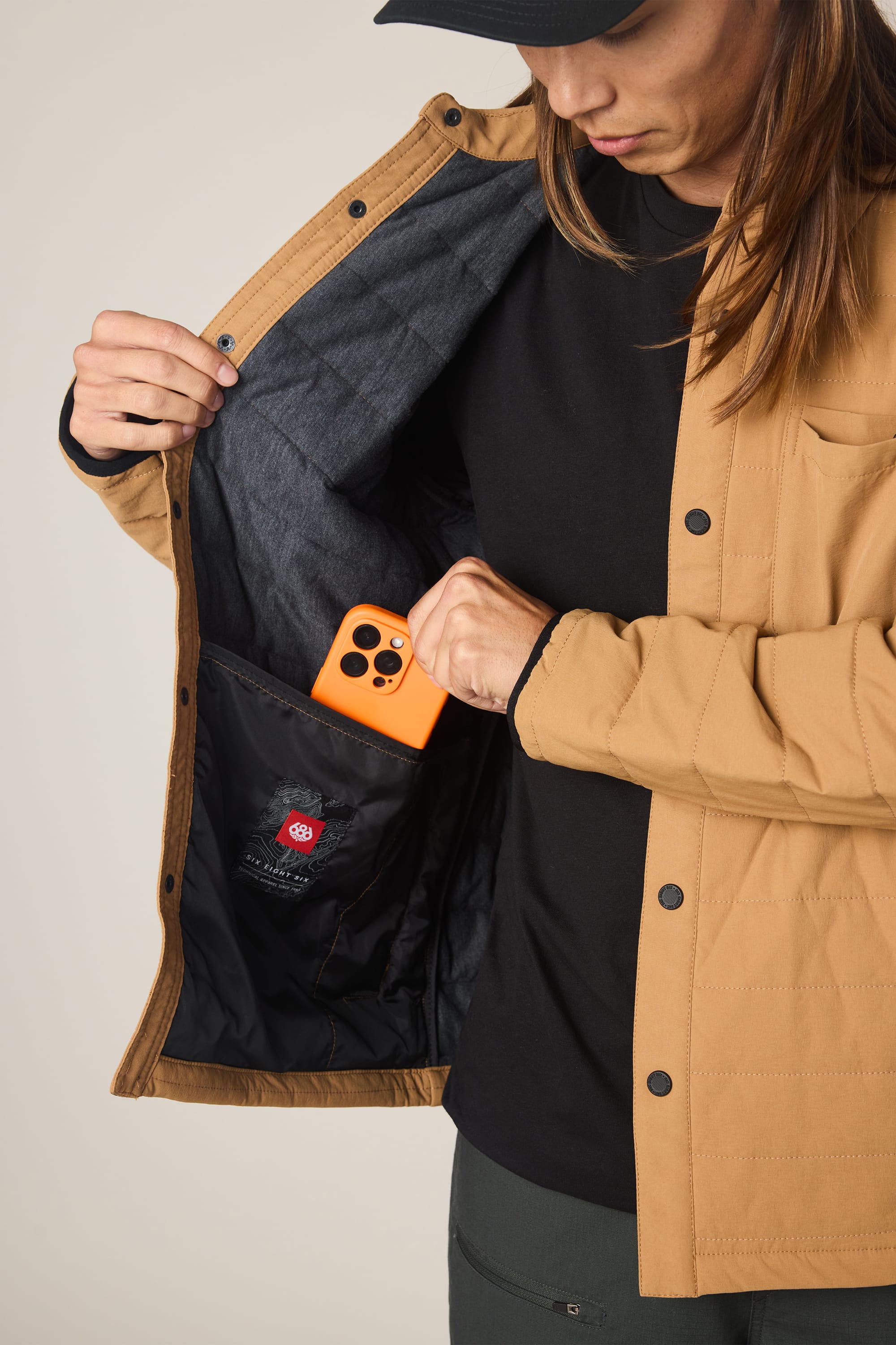 A person in a tan 686 Men's Thermaldry Insulated Shacket and black shirt places an orange smartphone into the shacket's inner pocket, exposing its lining and label. Their face is partly visible as they look down.