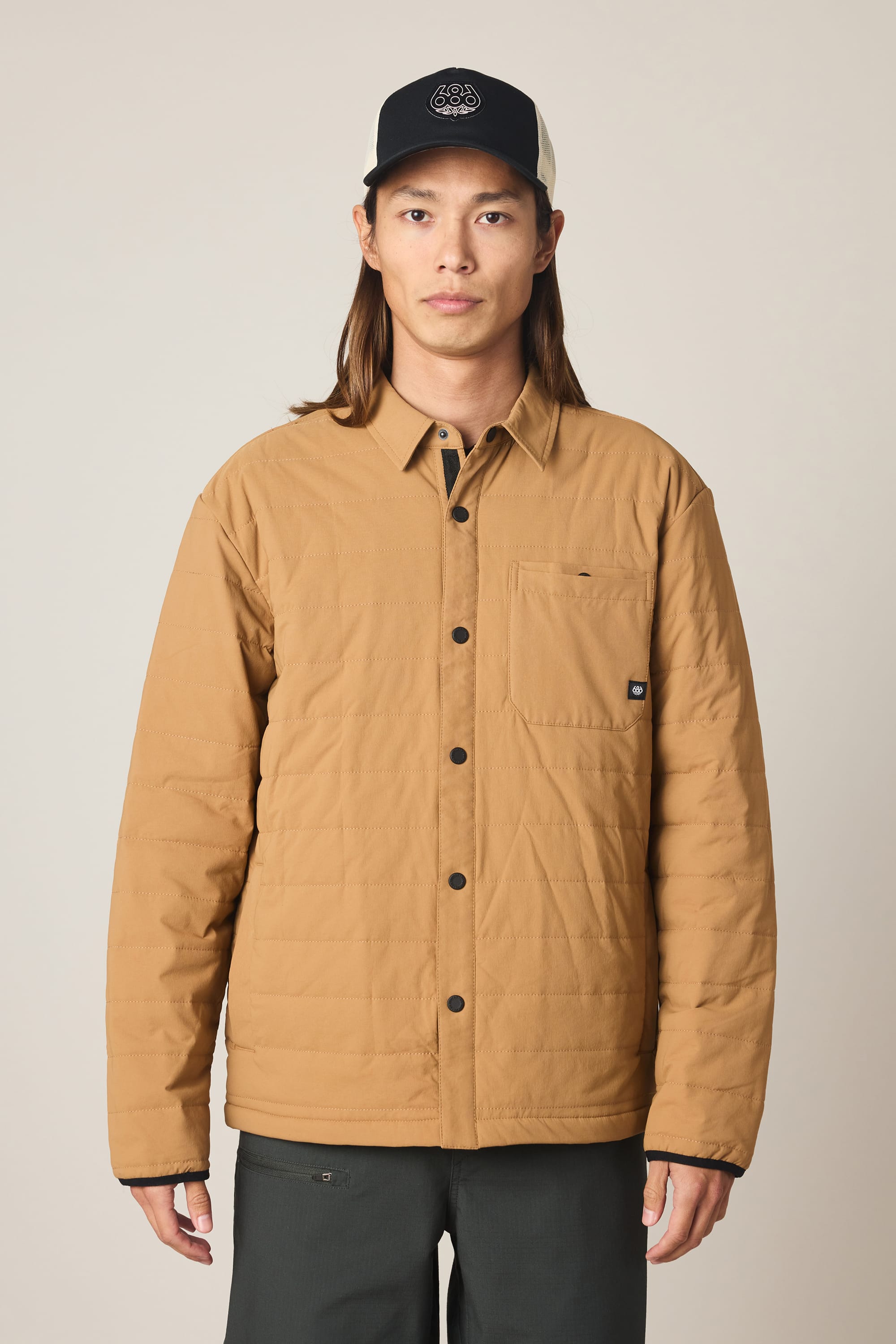A person wearing the 686 Men's Thermaldry™ Insulated Shacket in tan and a black cap stands facing forward against a plain light background.