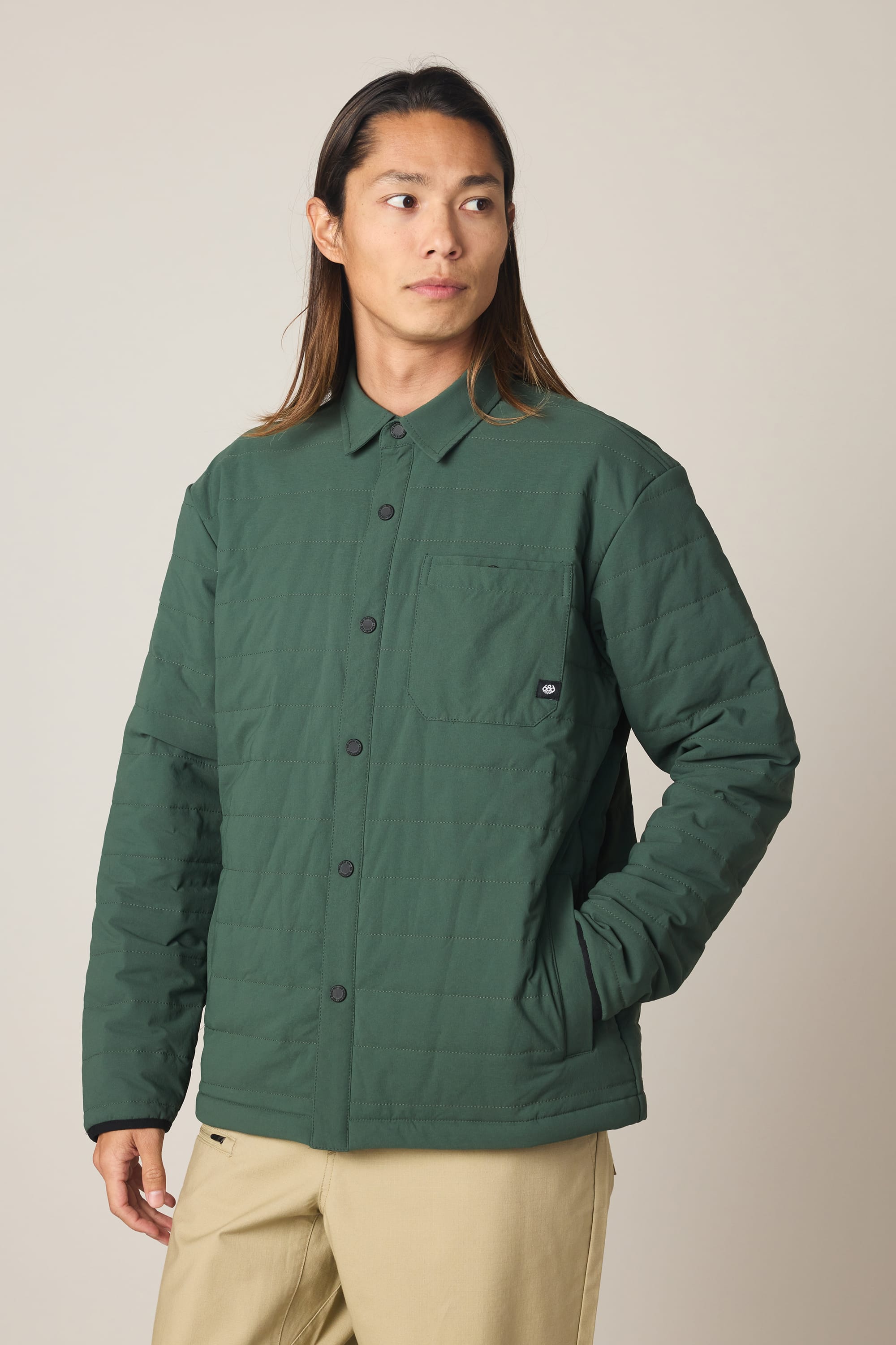 A person with long hair wears a forest green 686 Men's Thermaldry Insulated Shacket with front pockets and beige pants, standing against a plain light background, looking off to the side.
