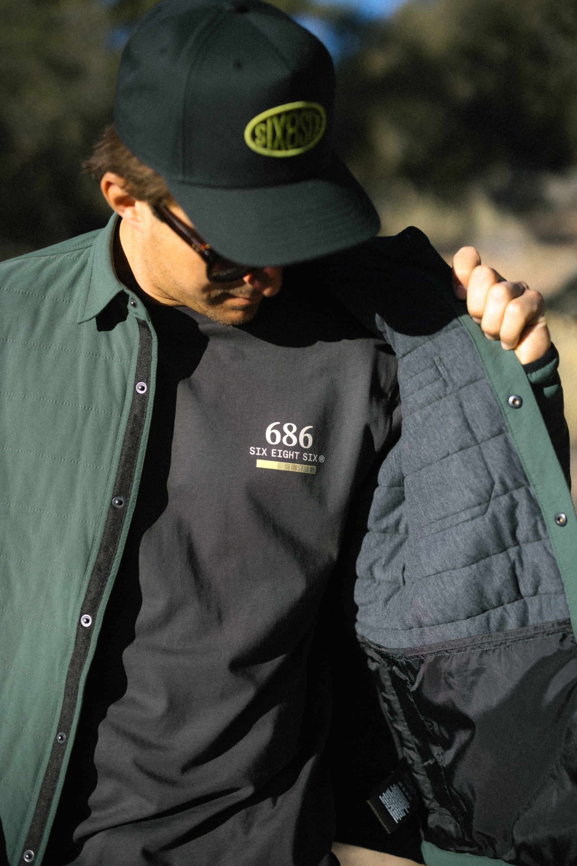 A person wearing sunglasses lifts their green 686 Men's Thermaldry™ Insulated Shacket to reveal a black T-shirt with "686 SIX EIGHT SIX" printed on it. They are also wearing a black cap with a yellow oval logo.