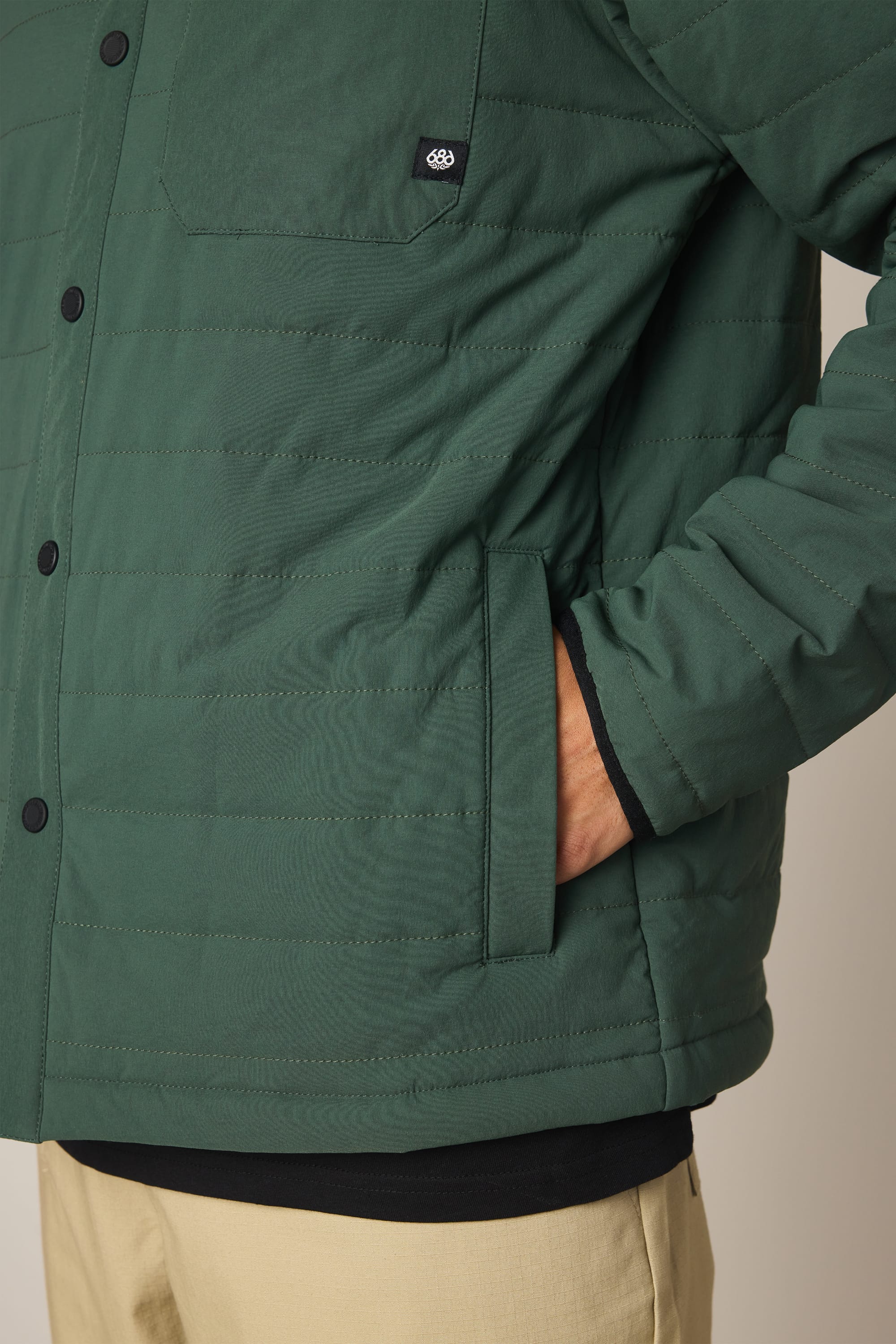 A person wears the 686 Men's Thermaldry Insulated Shacket in dark green with black buttons and tan pants. One hand is in the jacket pocket, and only the lower torso and part of one arm are visible.