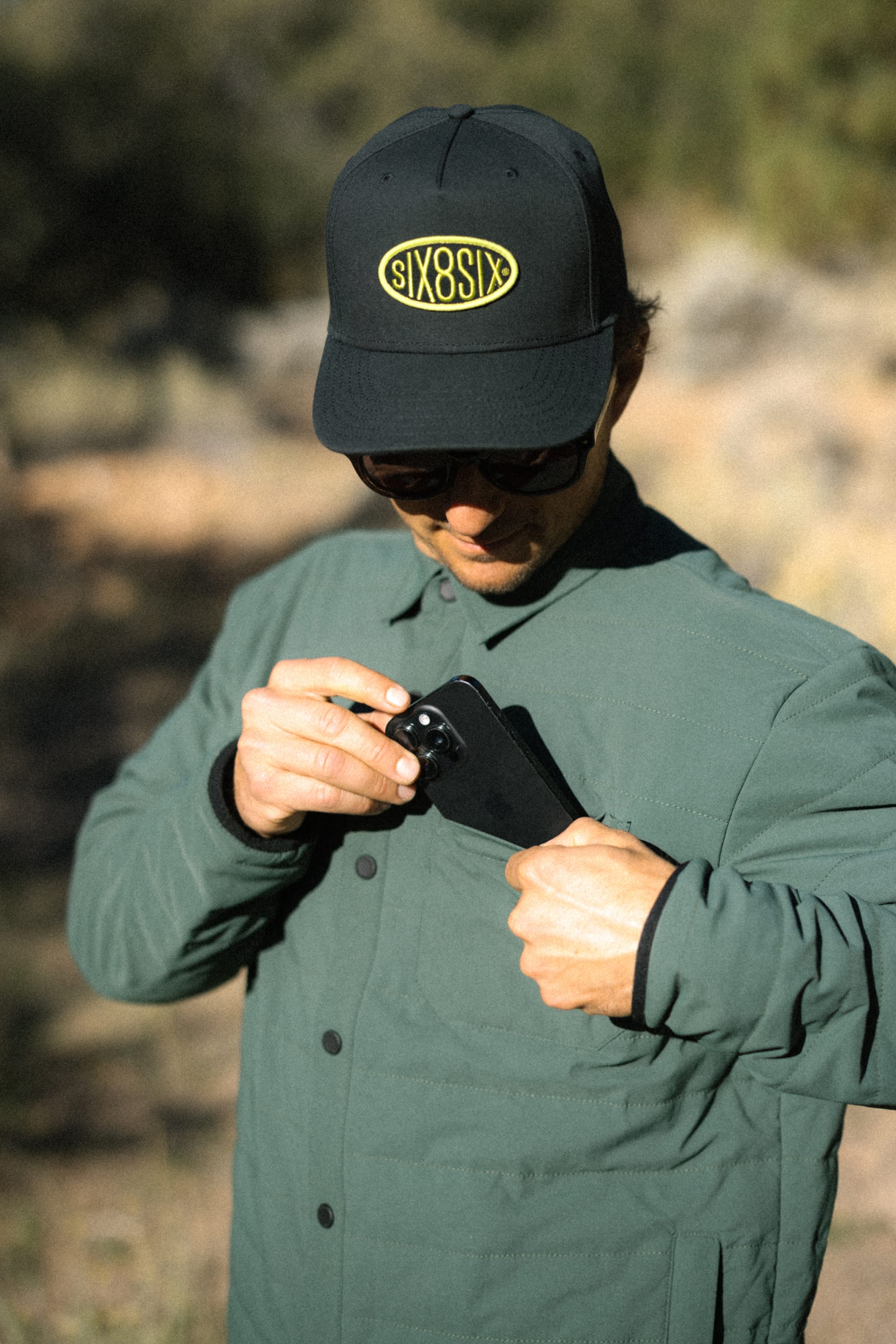 A person wearing a green 686 Men's Thermaldry™ Insulated Shacket and a black cap with “SIX O SIX” text is outdoors, looking down while placing a black smartphone into the shacket’s pocket.