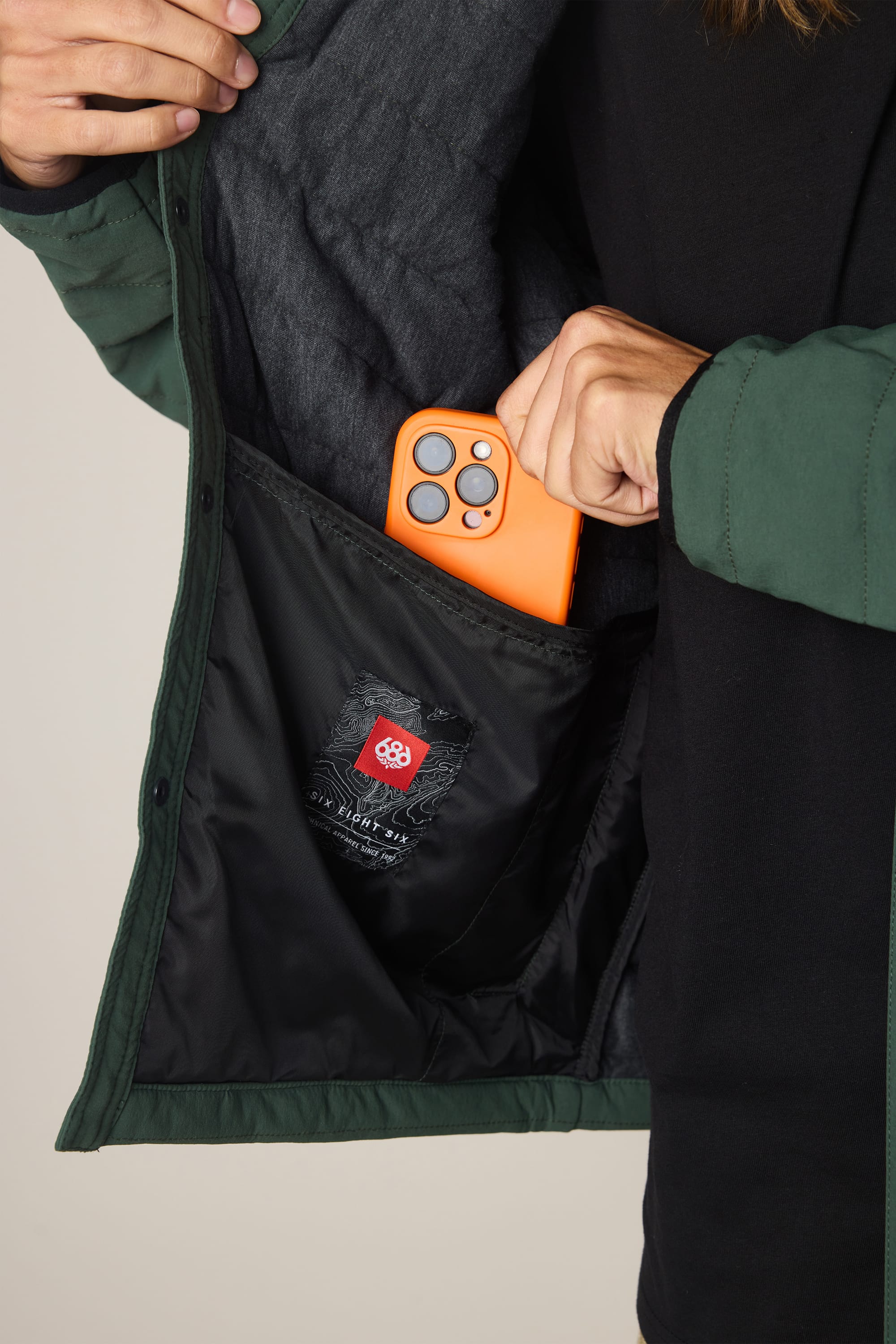 A person places an orange smartphone into the inner pocket of the 686 Men's Thermaldry™ Insulated Shacket, a green polyester jacket by 686 that features a black lining and a red logo patch inside.