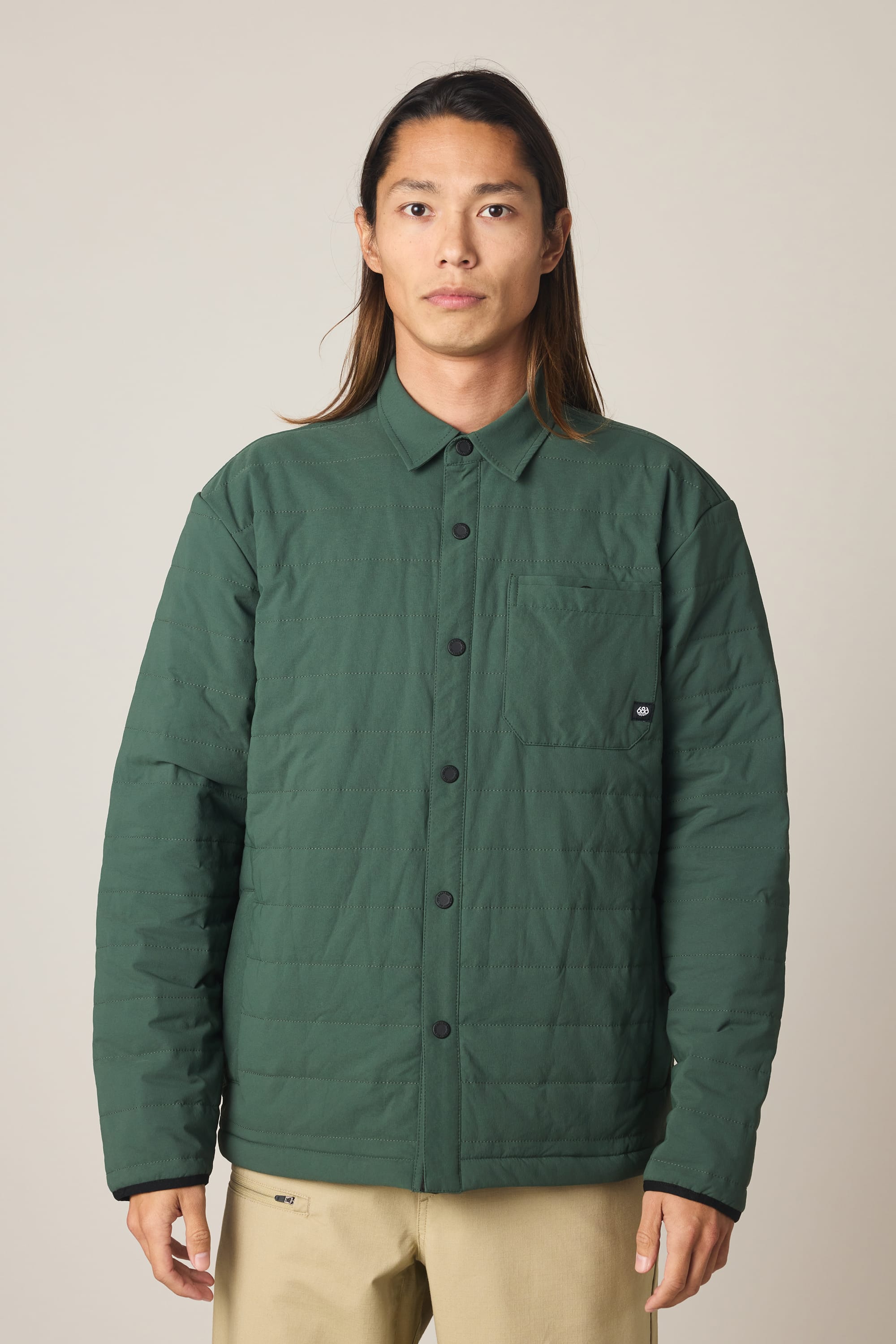 A person with long brown hair wears the 686 Men's Thermaldry Insulated Shacket in dark green and beige pants, standing against a plain light-colored background.