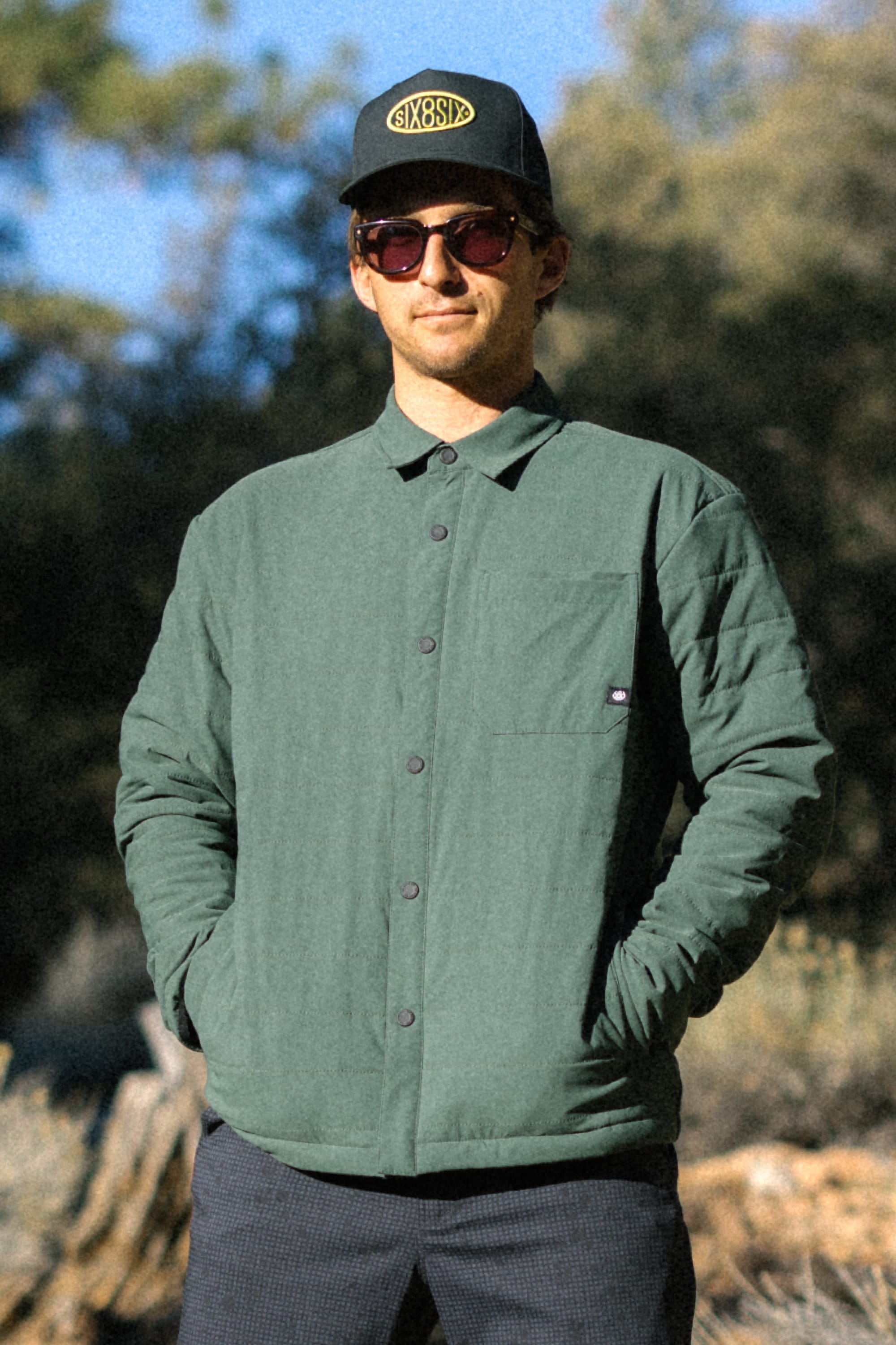A man wearing sunglasses, a dark cap, and the 686 Men's Thermaldry™ Insulated Shacket in green stands outdoors with hands in his pockets. Trees and sunlight fill the background.