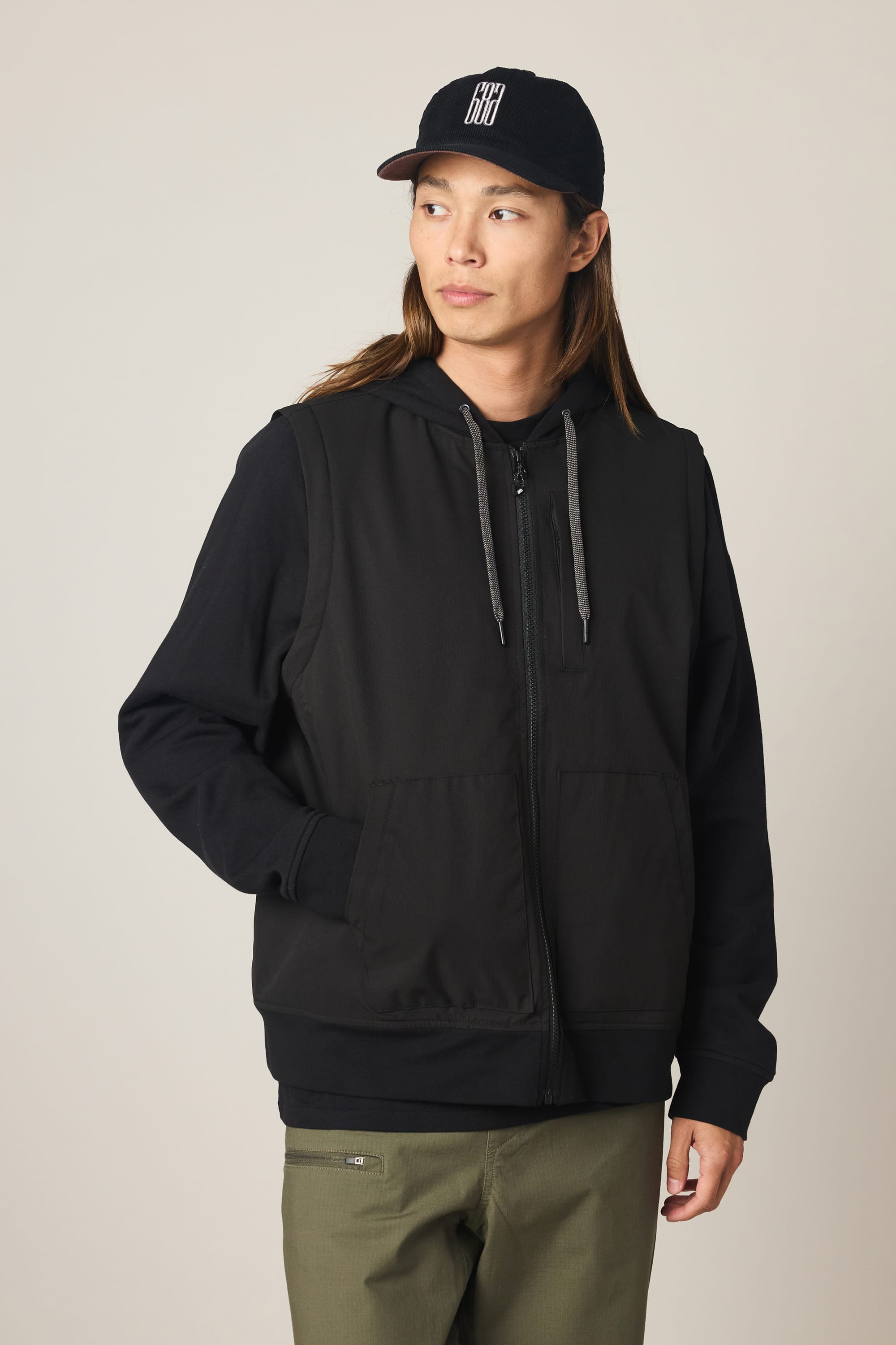 A person wearing a black cap, olive-green pants, and the 686 Men's Merge Quilted Hoody by 686—a black polyester jacket with gray drawstrings—stands against a plain light background, looking slightly to the side with hands in the jacket pockets.