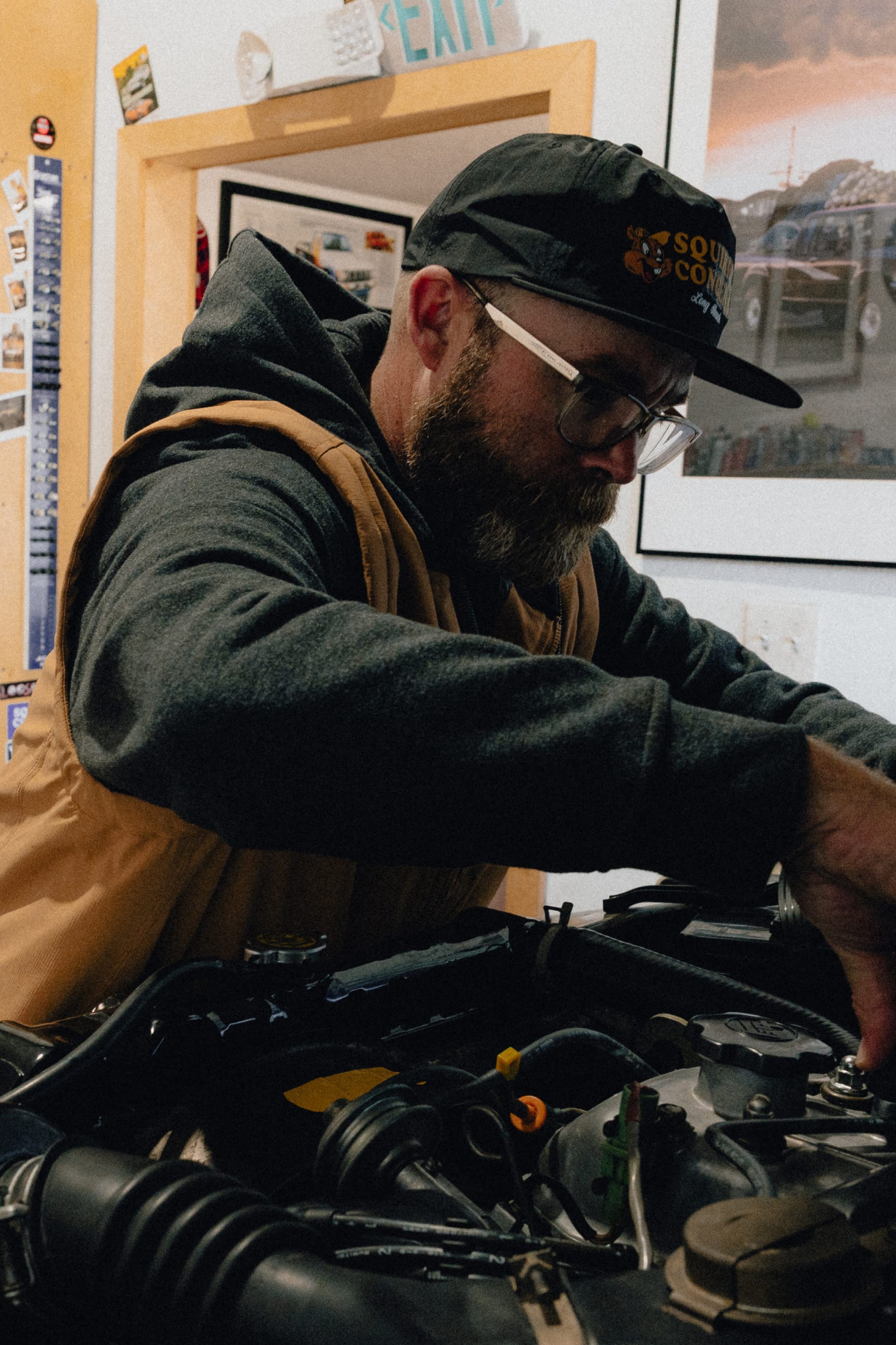 A bearded man wearing glasses, a cap, and a 686 Men's Merge Quilted Hoody works intently on a car engine in a garage surrounded by photos and various tools.