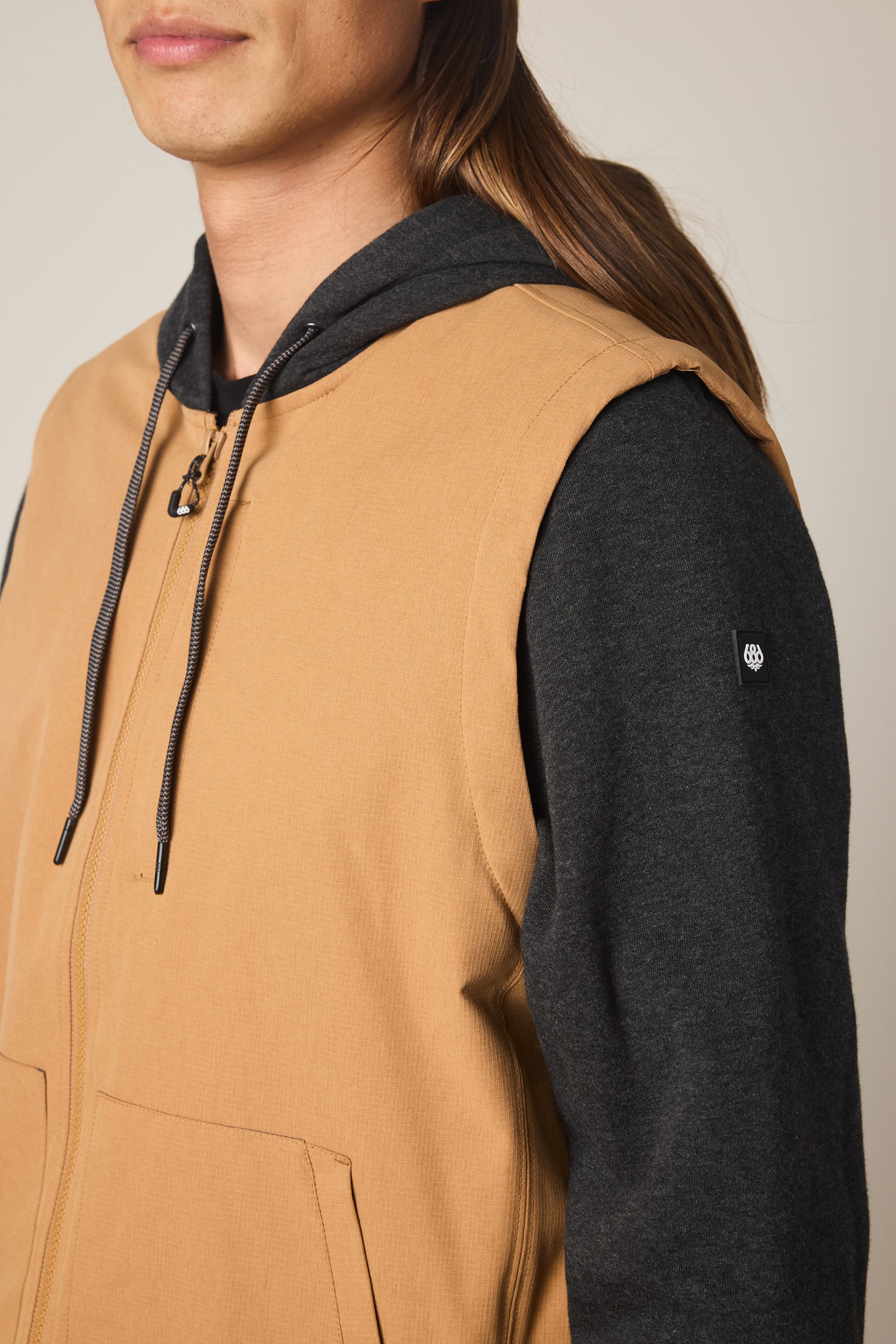 A person with long brown hair wears the 686 Men's Merge Quilted Hoody, a black polyester knit jacket featuring drawstrings and a small circular logo on the sleeve, under a tan sleeveless vest. Only the lower half of their face is visible.