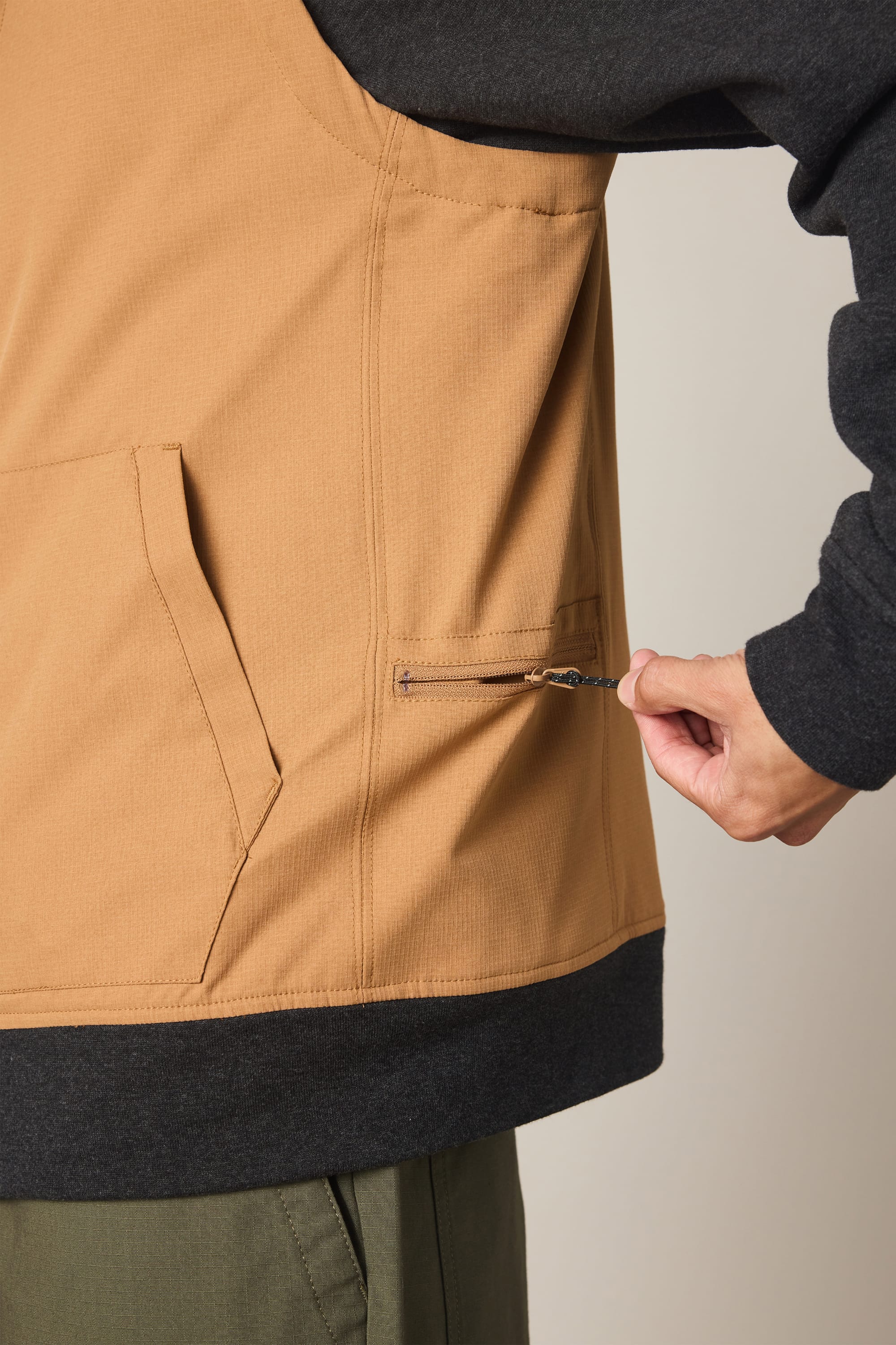 A person wearing the 686 Men's Merge Quilted Hoody in tan and black adjusts a small side zipper pocket; only their torso and hand are visible.