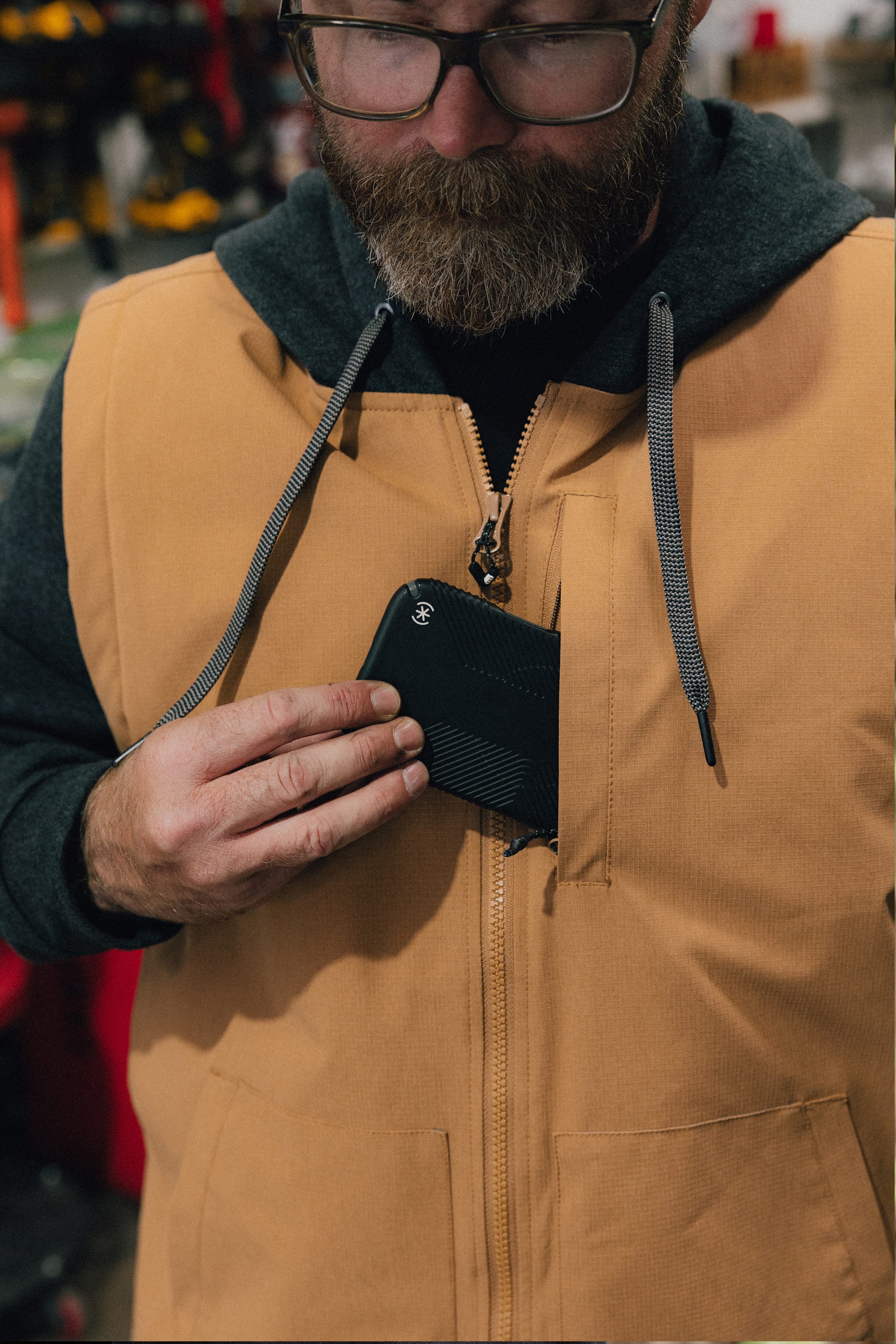 A bearded person wearing glasses and a tan 686 Men's Merge Quilted Hoody places a black zippered wallet into the chest pocket. The background is blurred, with tools and shelves visible.