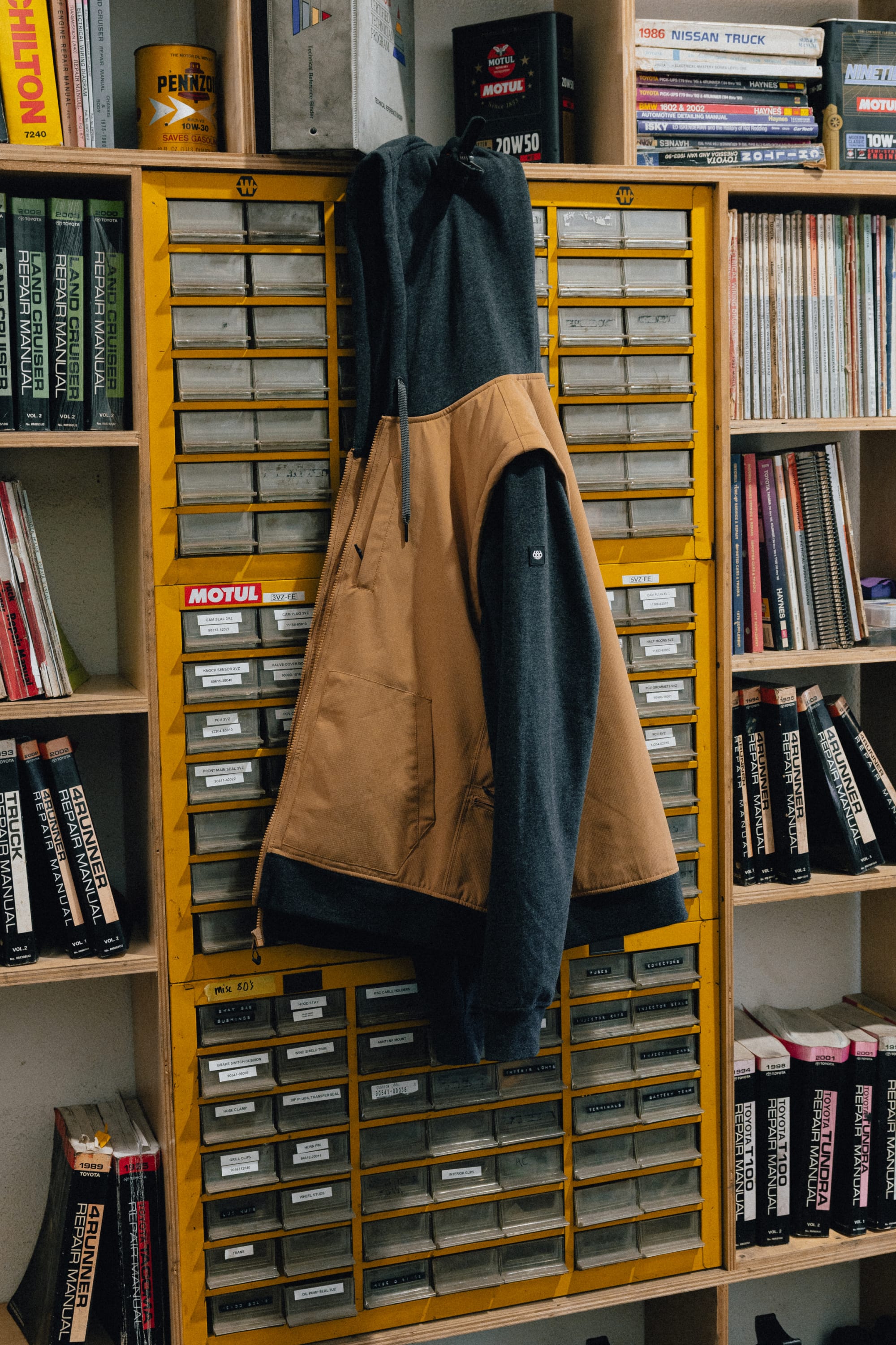 A 686 Men's Merge Quilted Hoody in tan and black hangs on a yellow cabinet with many small drawers, surrounded by bookshelves filled with books and magazines.
