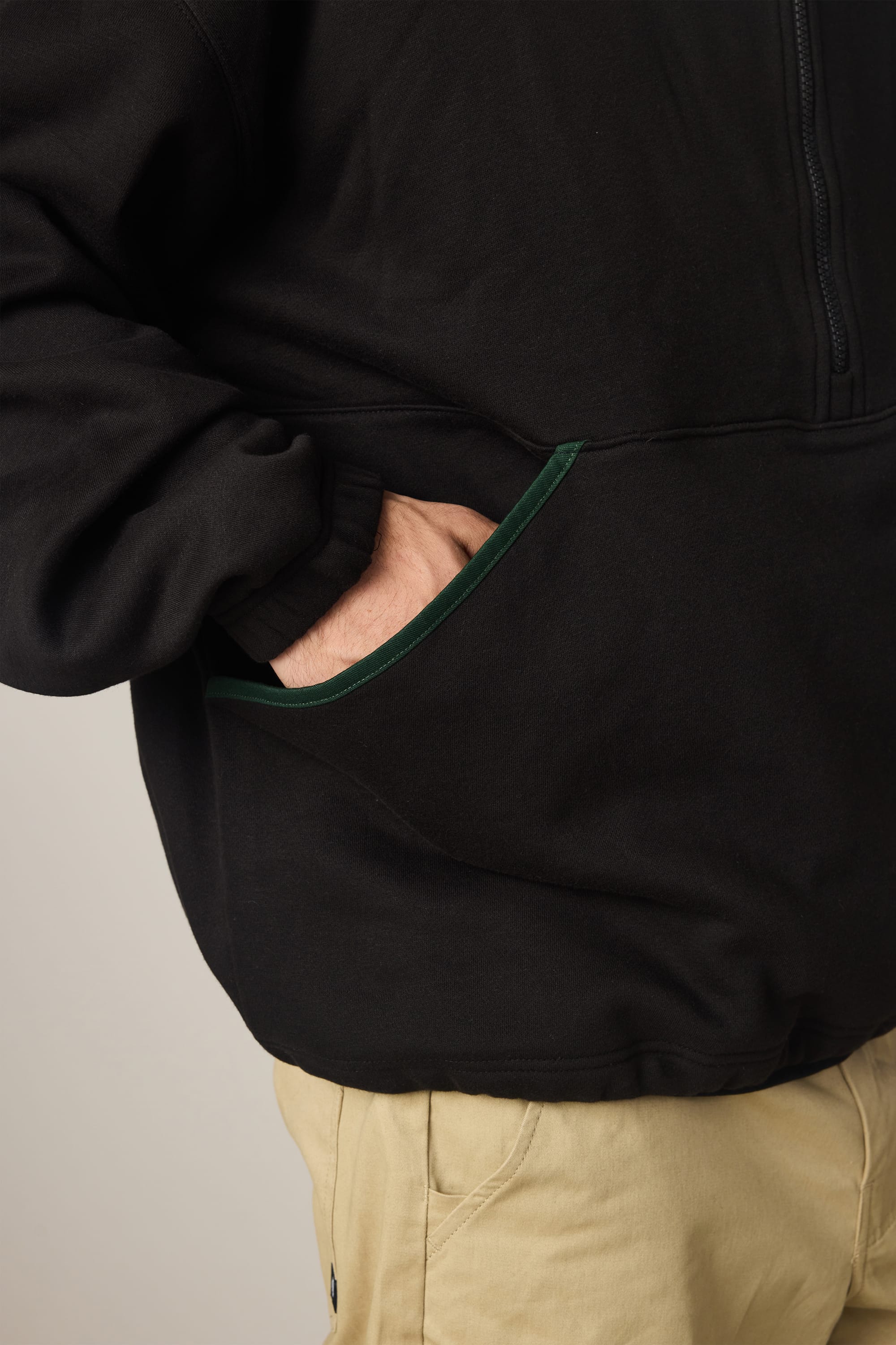 A person wearing the 686 Men's Burnout Anorak—a black, waterproof jacket with a green-trimmed pocket—and beige pants. Only the torso and upper legs are visible, with one hand in the pocket.