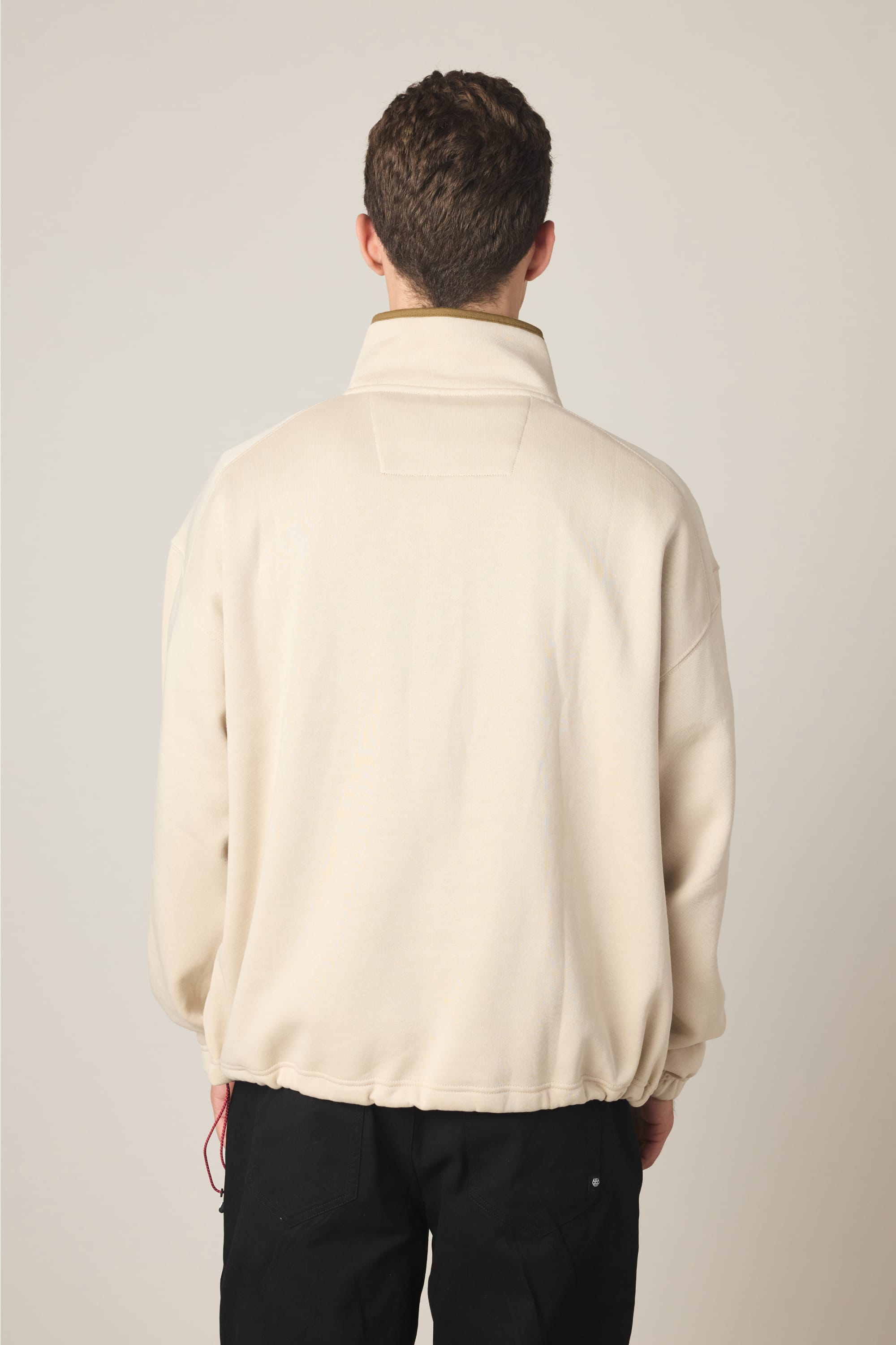 A person with short brown hair is standing with their back to the camera, wearing the 686 Men's Burnout Anorak in beige and black pants against a plain light background.