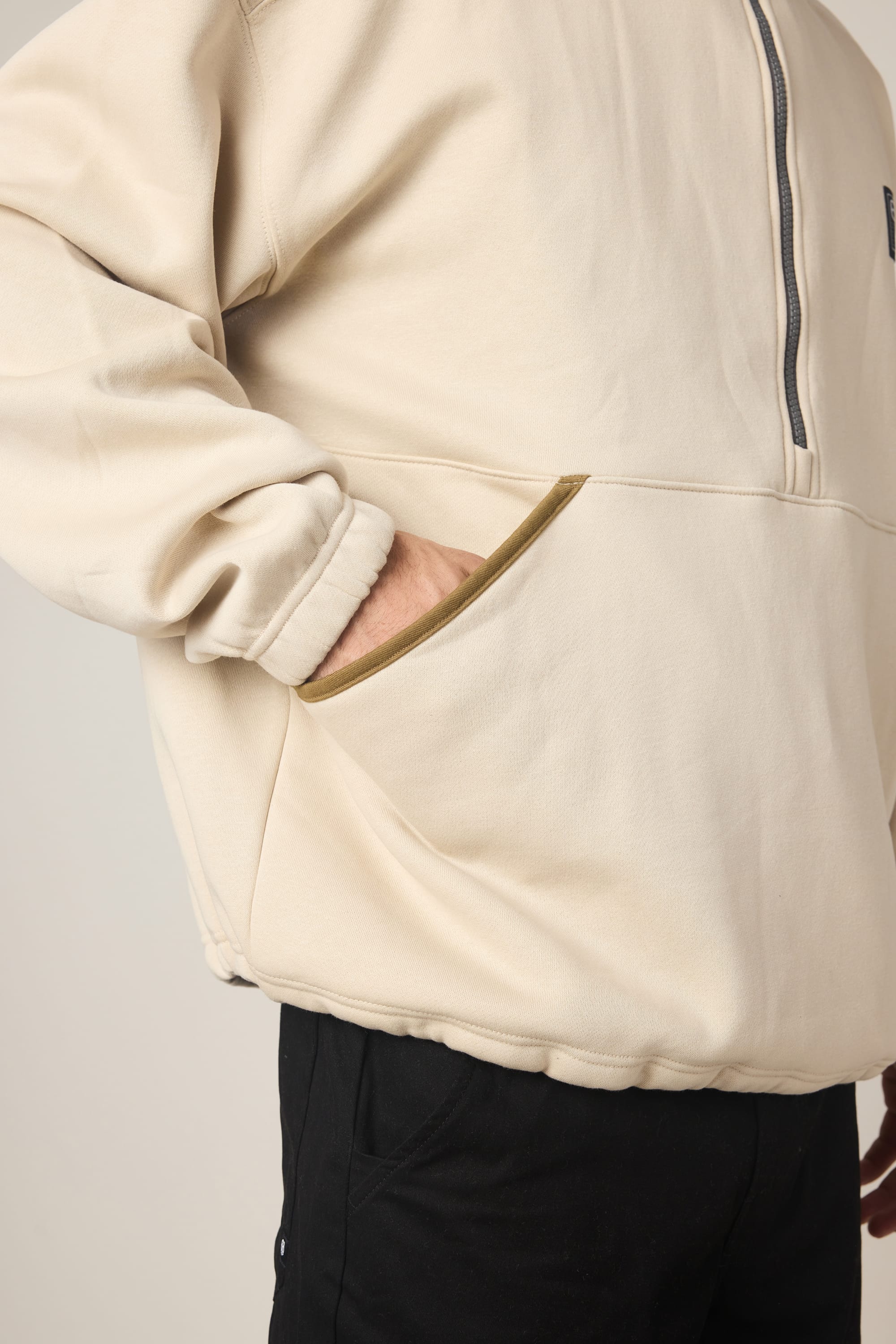 A person wearing the 686 Men's Burnout Anorak—a beige woven jacket with a zip-up design—has one hand in the pocket and is paired with black pants. The image is cropped to show only the torso and upper legs.