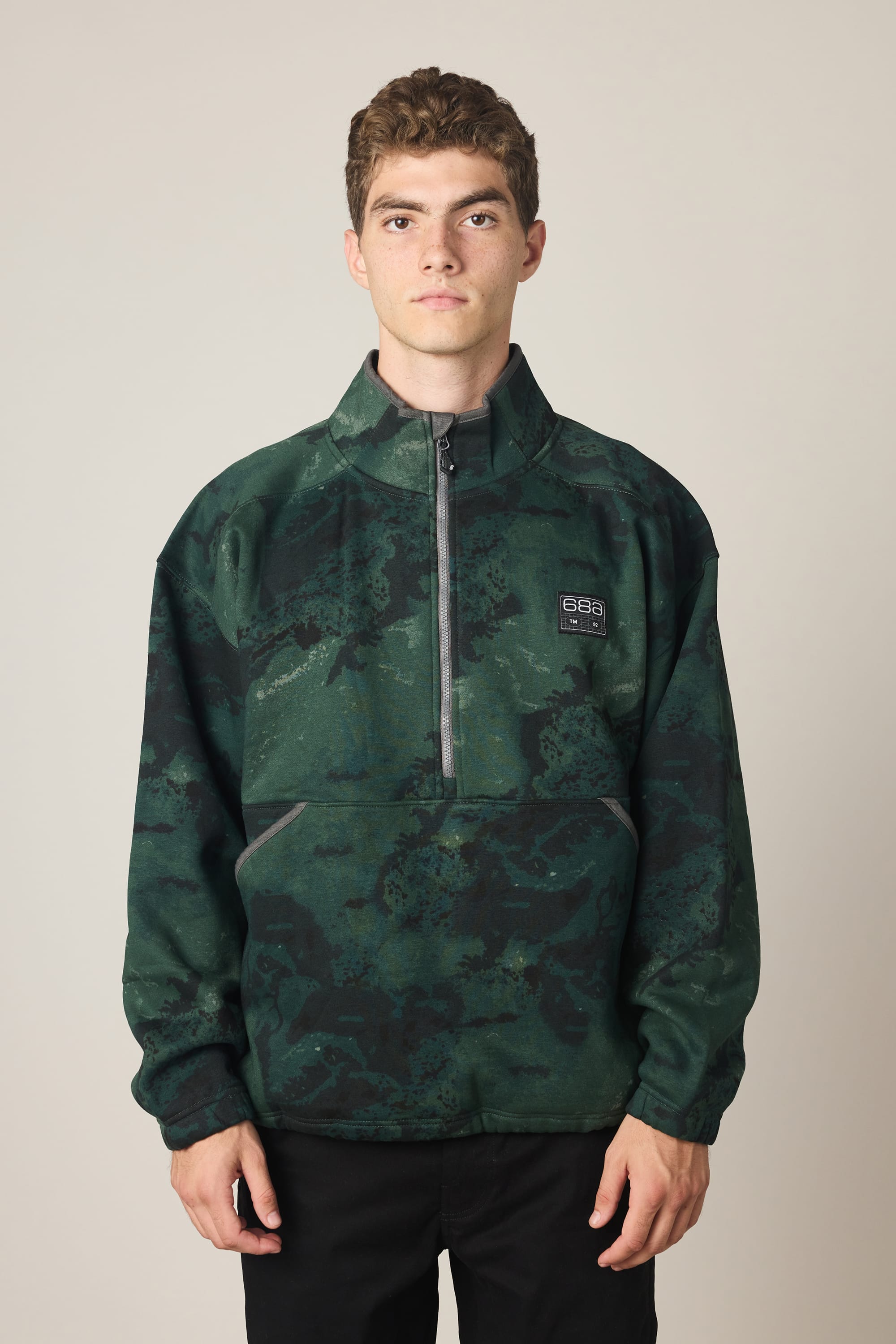 A young man with short brown hair wears the 686 Men's Burnout Anorak, a dark green camouflage half-zip jacket with a small 686 logo on the chest, standing against a plain light background.