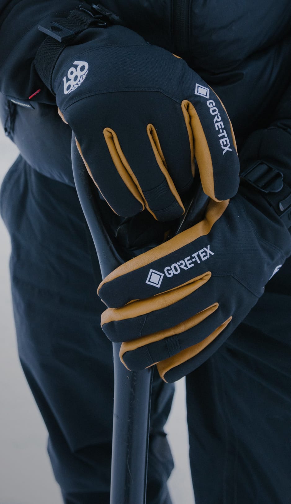 A person wearing black and yellow GORE-TEX gloves holds the handle of a ski pole or tool, dressed in dark winter clothing, with a snowy background visible.