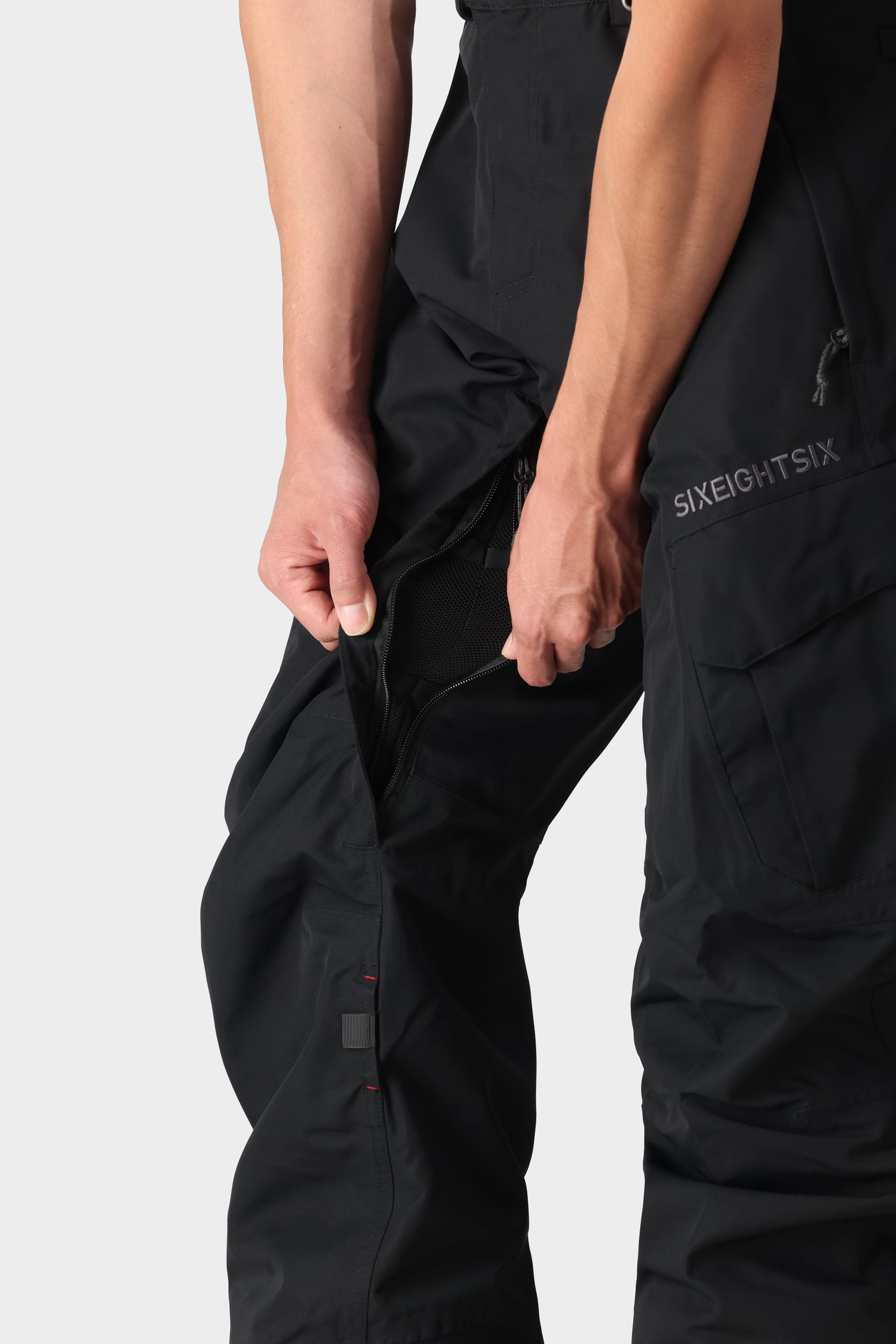 A person unzips a black pant leg of the 686 Men’s SMARTY® 3-in-1 Cargo Pant to reveal a mesh lining. The pants feature the 686 logo printed near the pocket, with only the waist down and part of the arms visible.