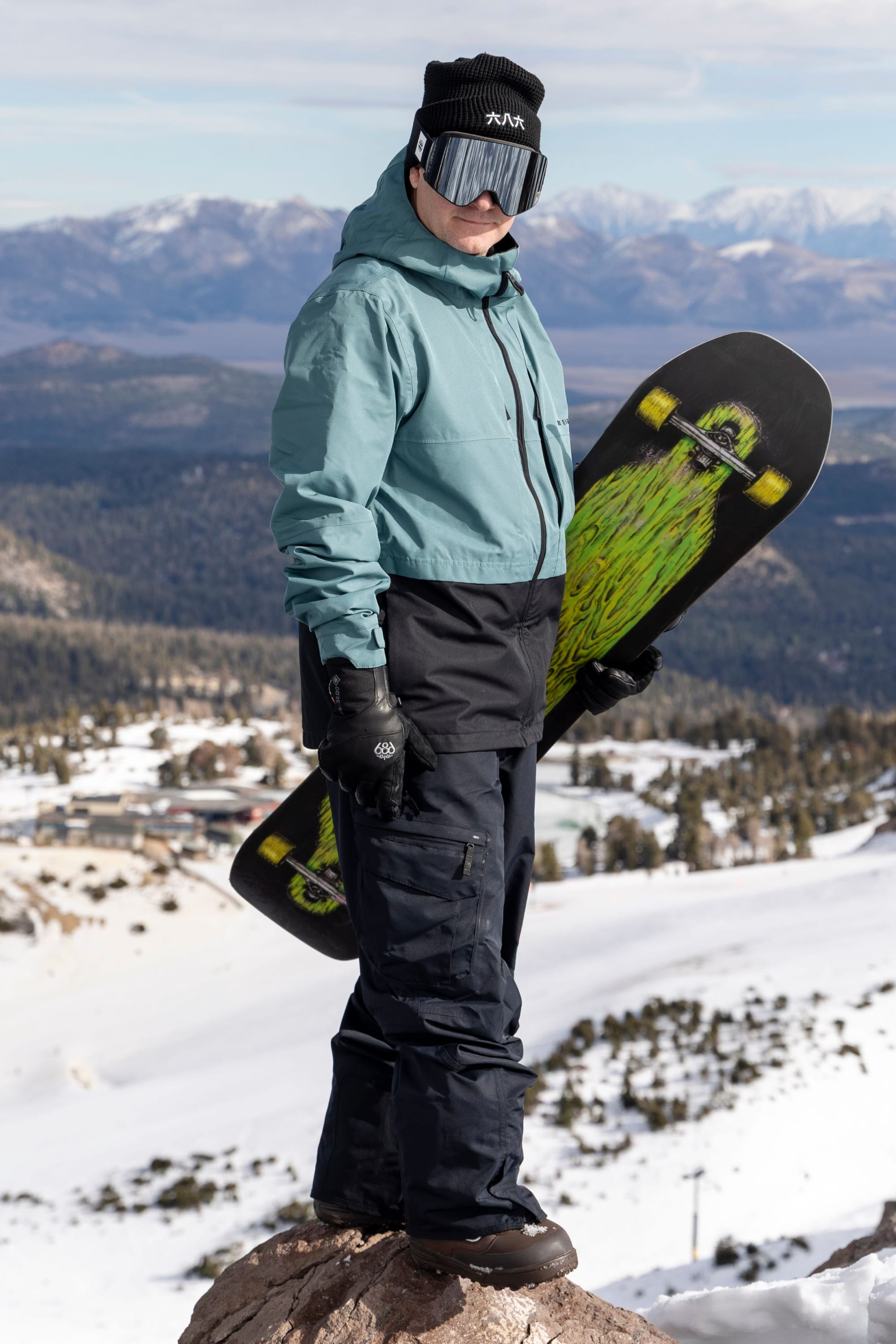 A person dressed in 686 Men’s SMARTY® 3-in-1 Cargo Pant winter gear, complete with goggles and a beanie, stands on a snowy hill holding a snowboard with a green graphic. Snow-covered mountains and trees are visible in the background.