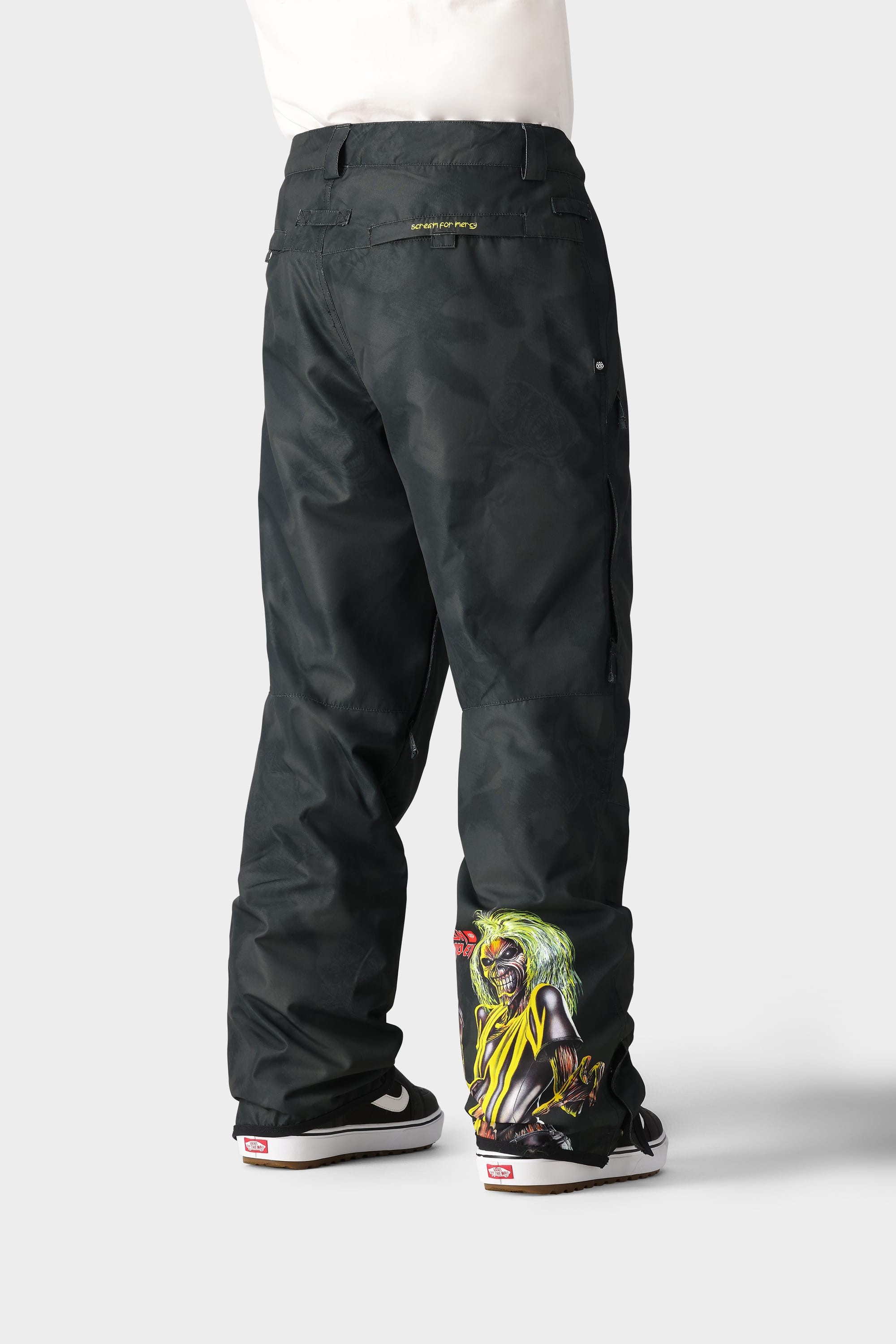 A person stands facing away in 686 Men's Standard Shell Pant by 686, featuring a colorful menacing clown graphic on the lower leg, secure pockets, and paired with black and white sneakers with red labels, set against a plain light gray background.