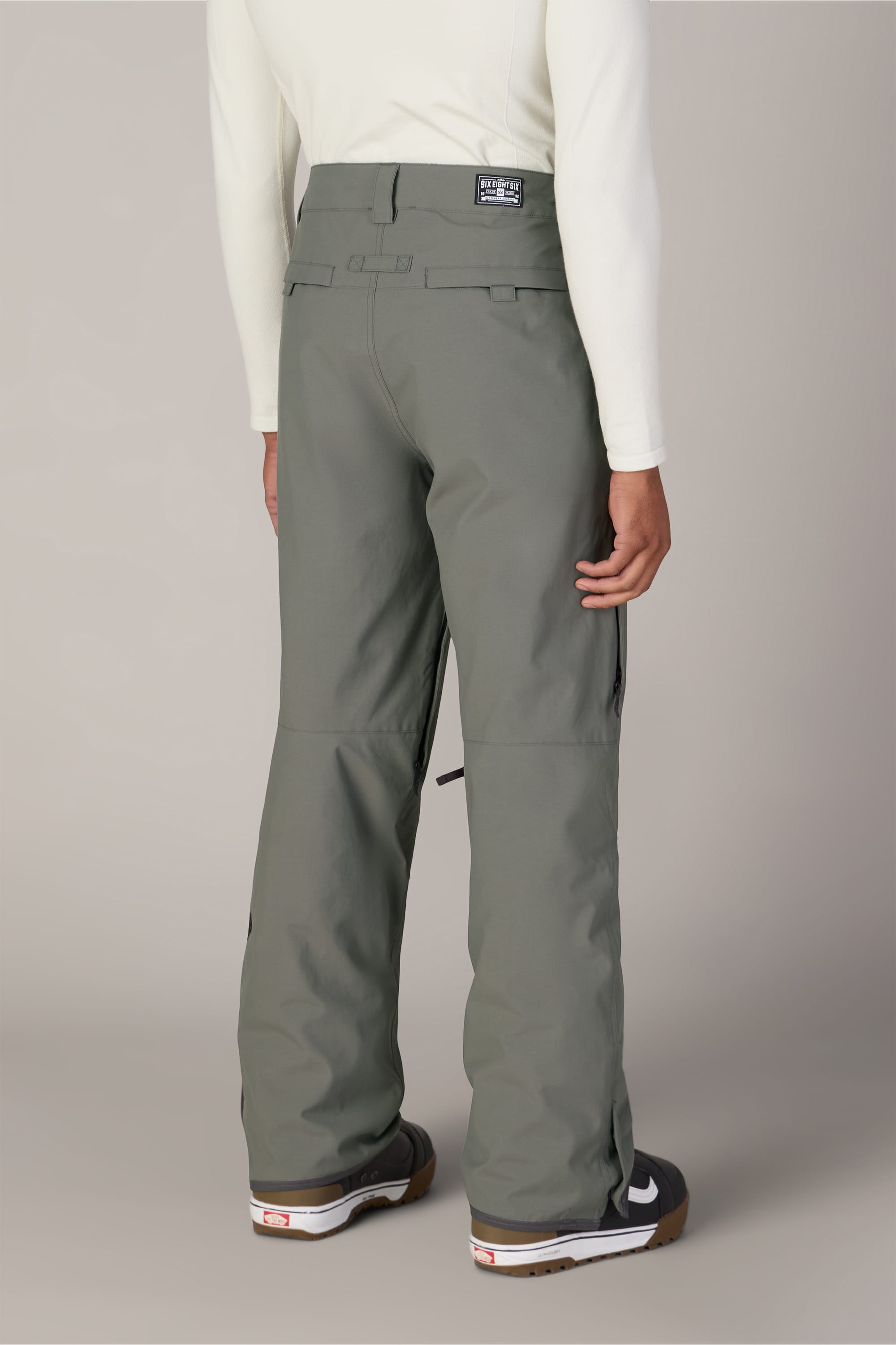 A person is standing facing away, showcasing the back details of the 686 Men’s Standard Shell Pant in affordable sage green, paired with a white long-sleeve shirt and black-and-white shoes. The waterproof pants by 686 are clearly visible.