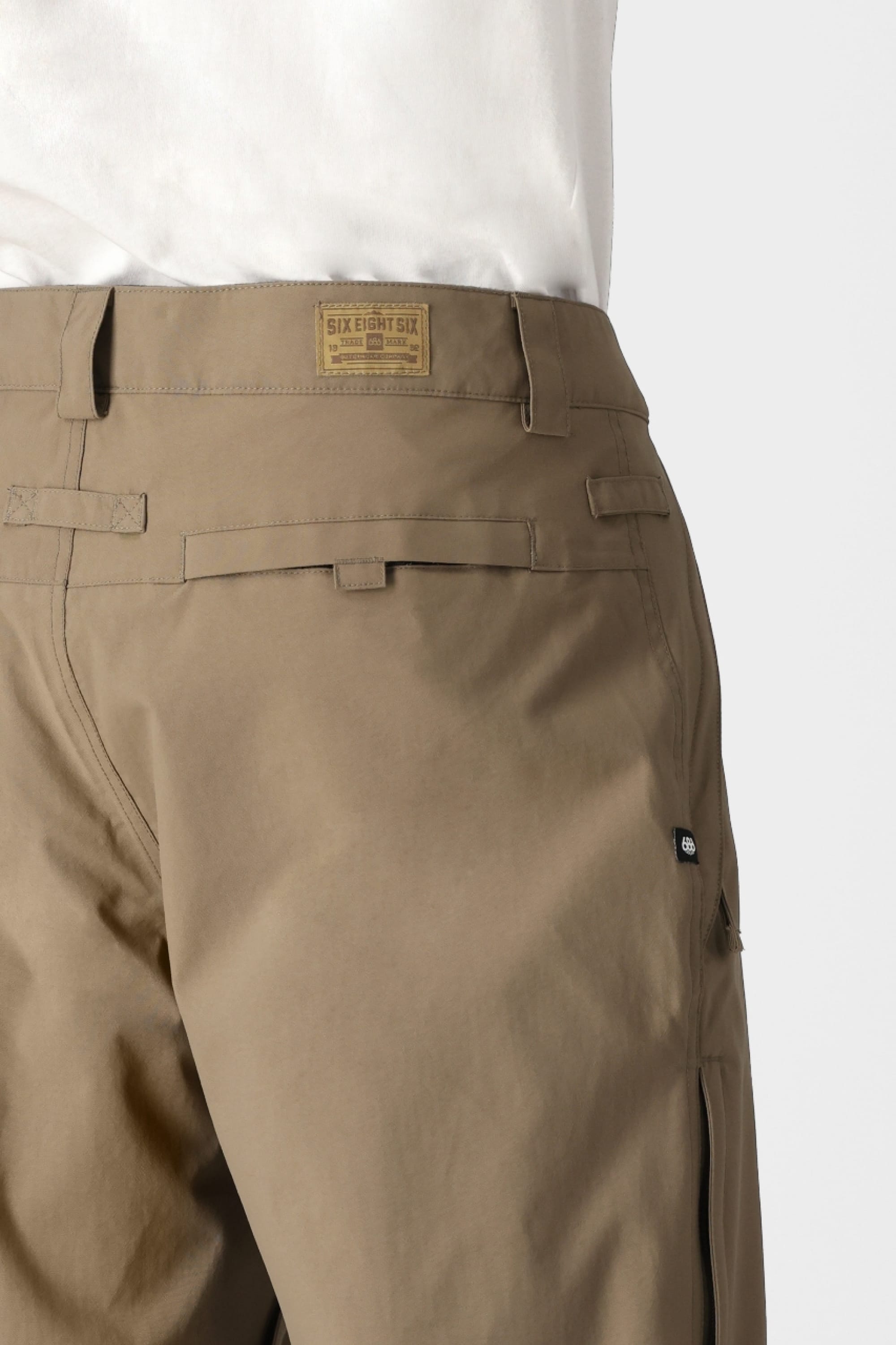 A person is wearing the 686 Men’s Standard Shell Pant by 686, featuring visible belt loops, a black button, and a tan 686 label on the waistband. The white shirt is tucked in above the waistband, and the background is plain light gray. These affordable pants offer a stylish look with their waterproof design for added functionality.