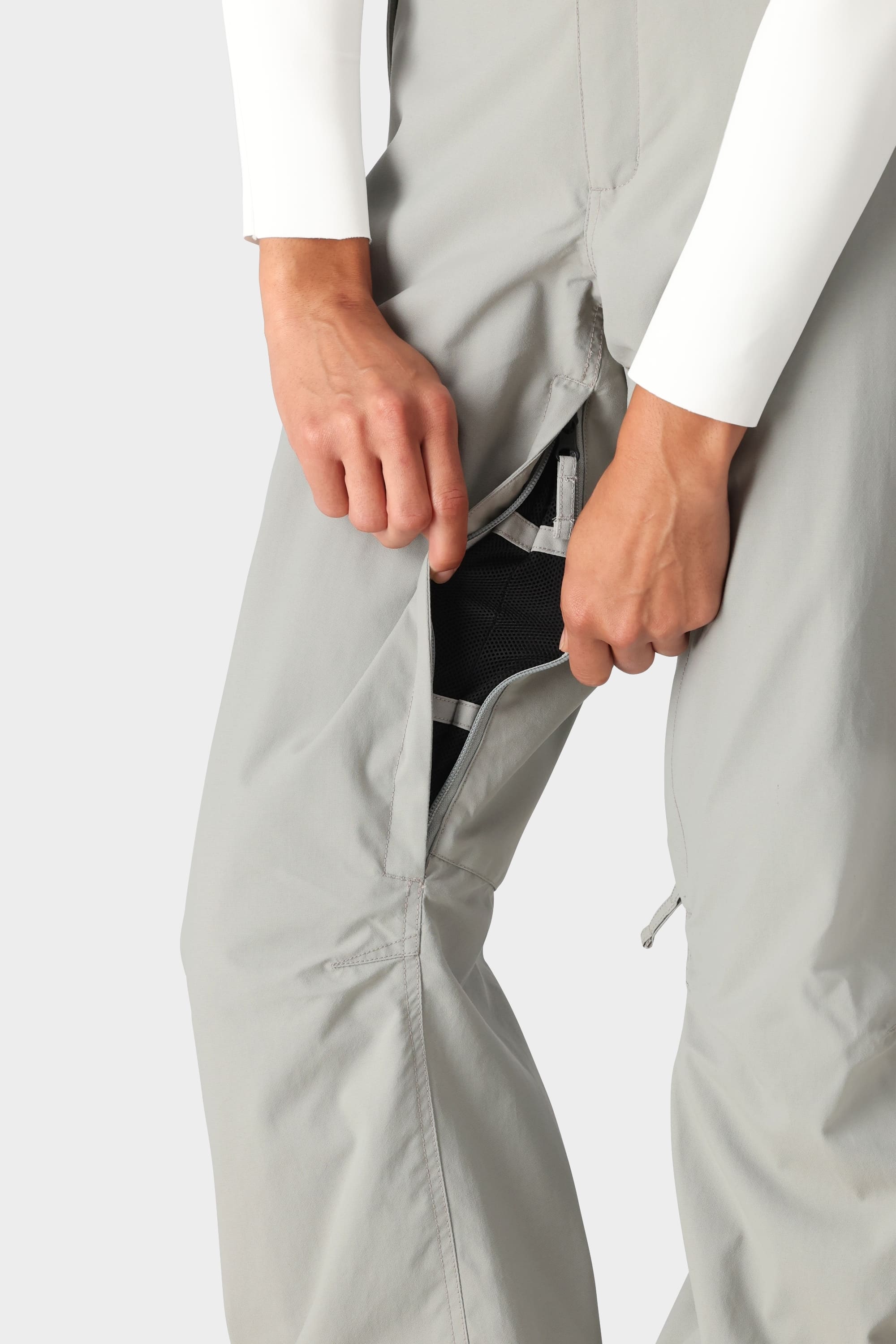 A person in 686 Womens Standard Shell Pant in light gray and a white long-sleeve shirt unzips a vertical thigh zipper, revealing black mesh lining.