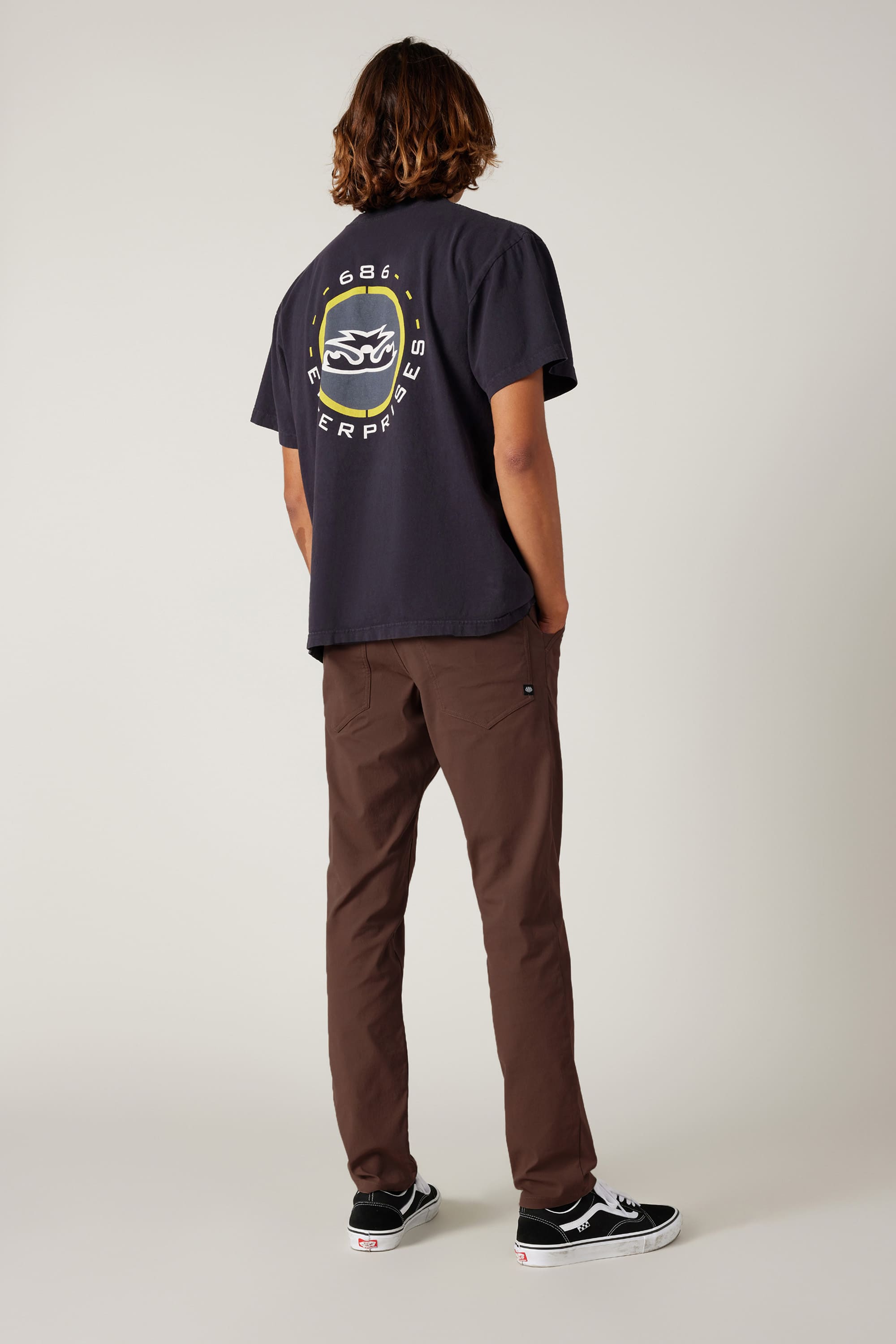 A person with shoulder-length hair faces away, wearing a black tee with a circular back graphic, 686 Men's Everywhere® Pant - Slim Fit in stain-resistant brown, and black-and-white sneakers against a plain light background.