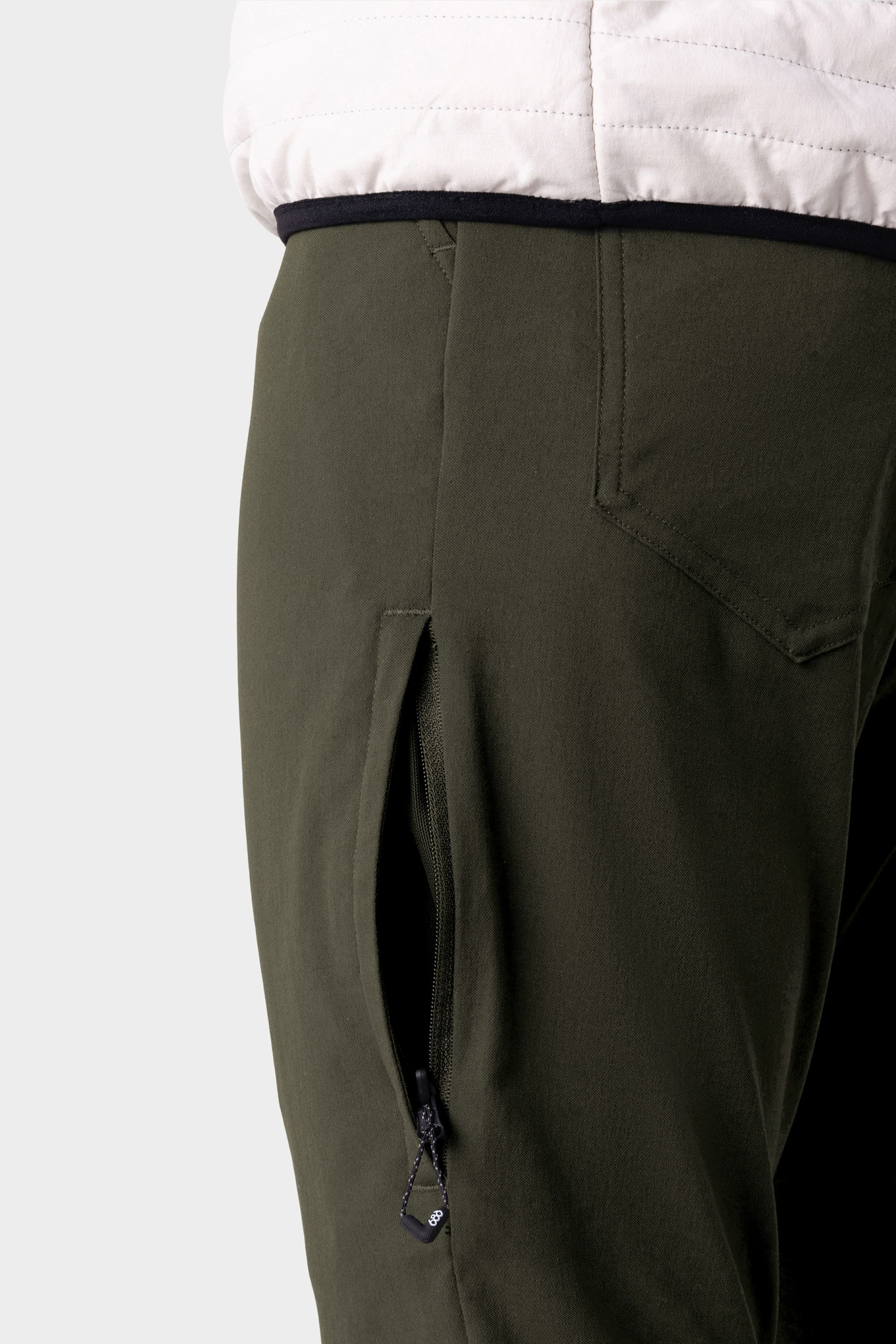 Close-up of a person wearing 686 Men's Everywhere® Pant - Slim Fit in olive green with a visible black zipper pocket, paired with a white quilted jacket.